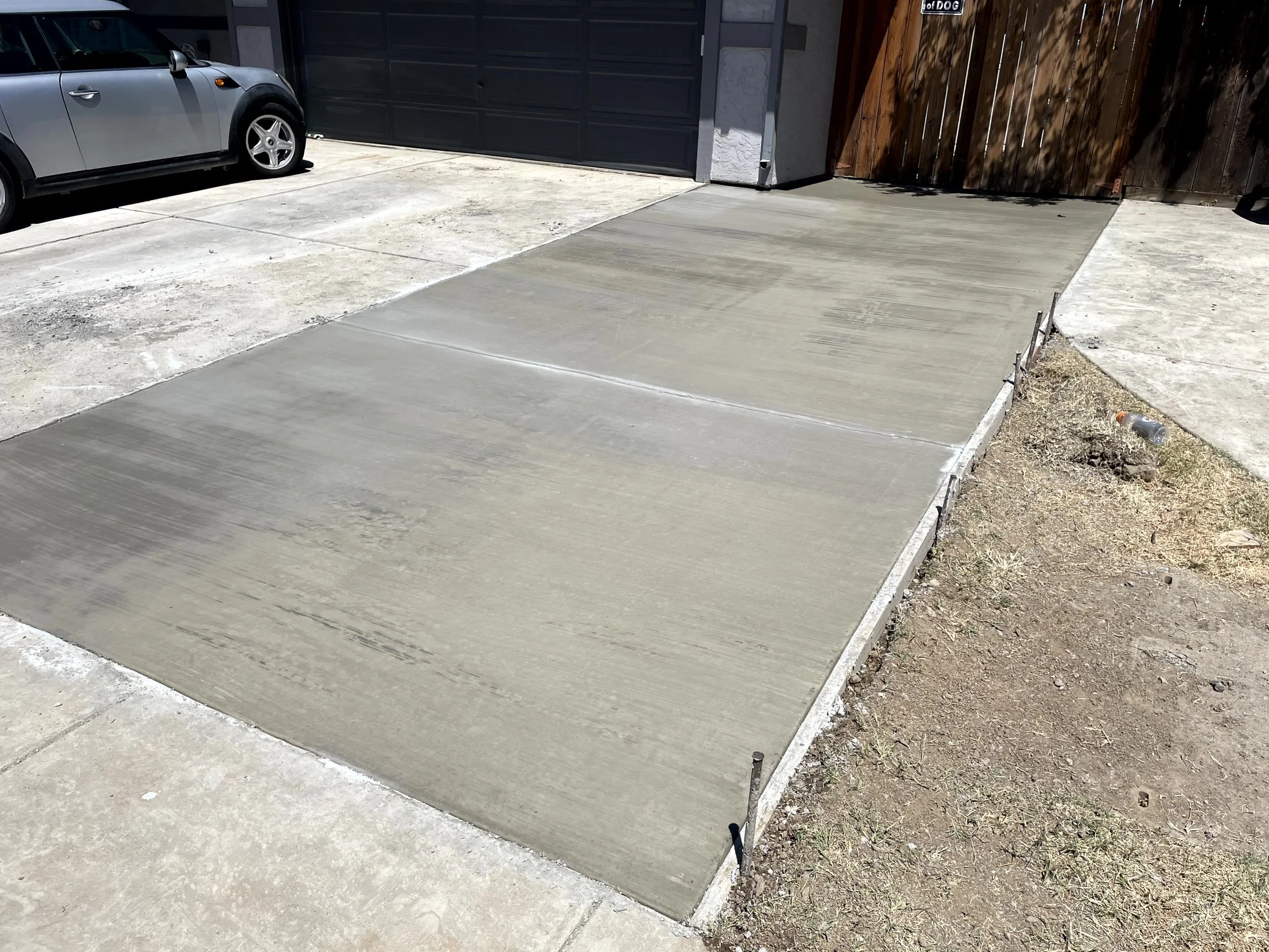 Freshly poured concrete driveway in front of a garage with a wooden gate, a gray car parked on the sidewalk, and a dirt area with a plastic bottle to the side.