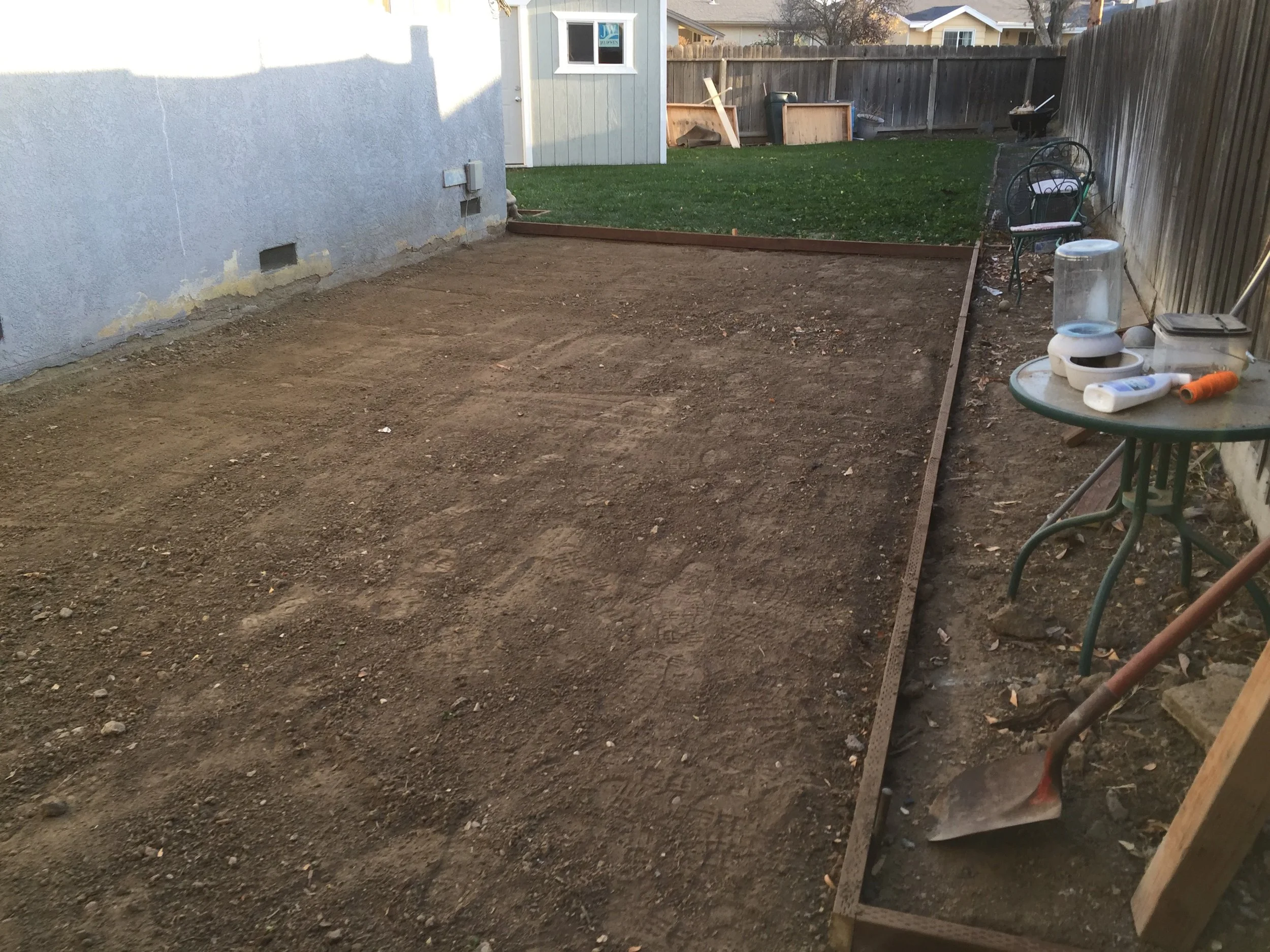 Backyard with a new dirt area bordered by wooden framing, adjacent to a grassy patch, with gardening tools and supplies on a small table to the right, and a green bench and fence in the background.
