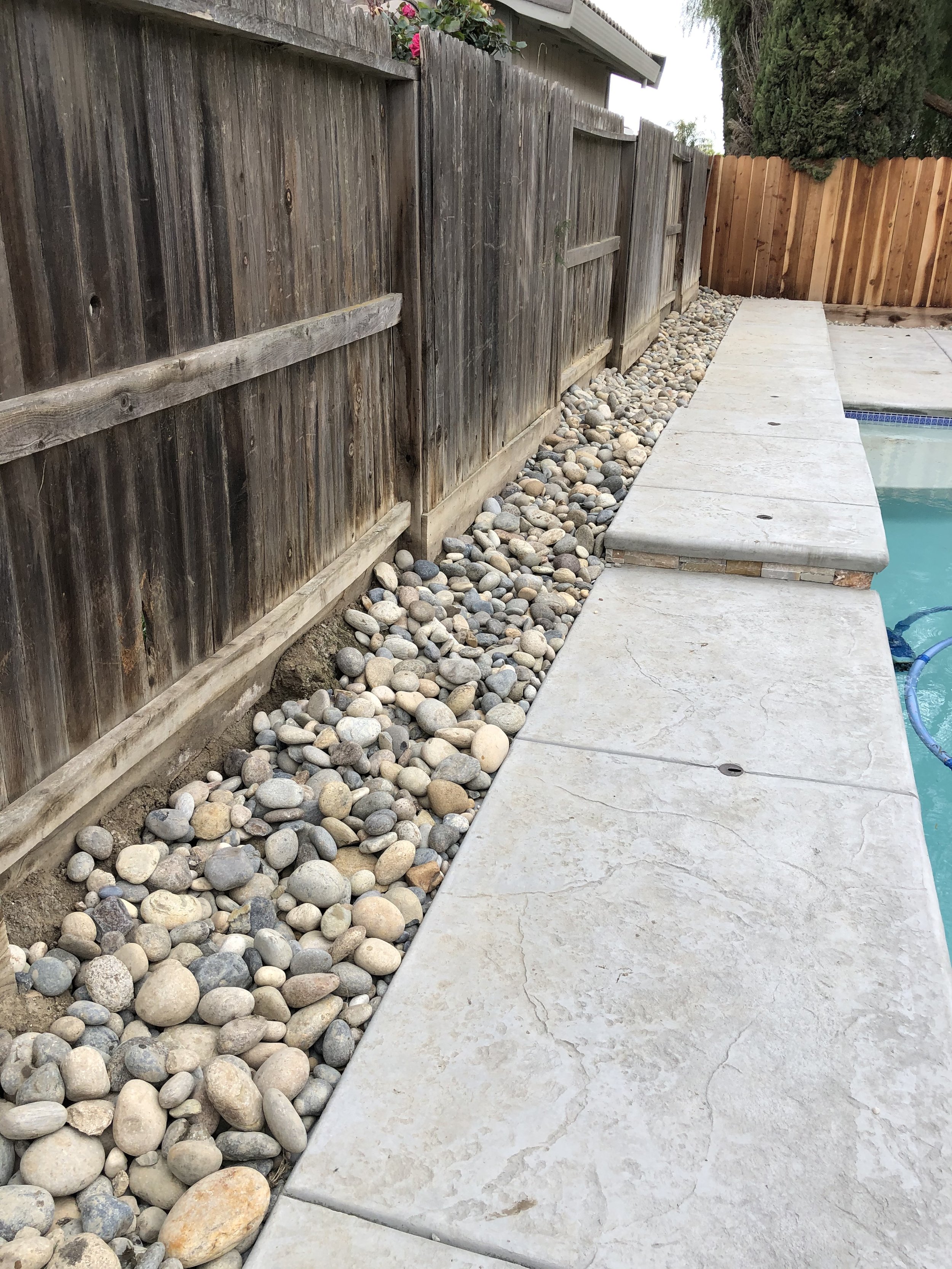 Concrete pool deck with a barrier of decorative rocks next to a wooden fence.
