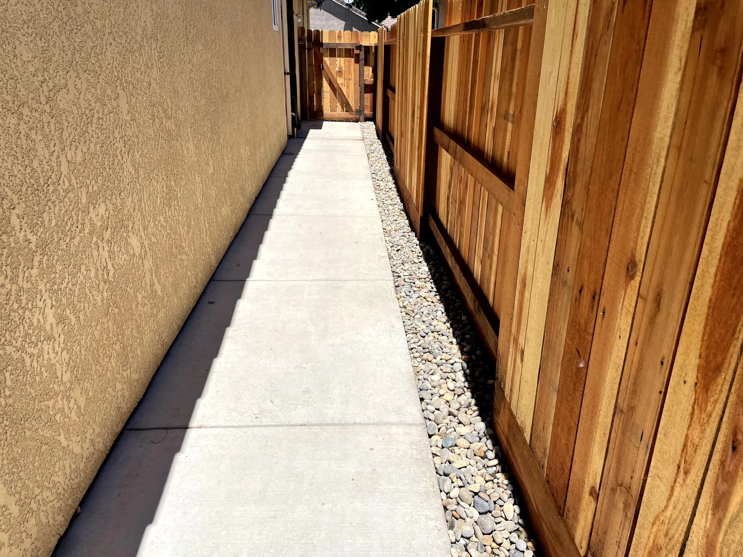 Concrete walkway between a yellow stucco wall and a wooden fence with small rocks lining the base of the fence.