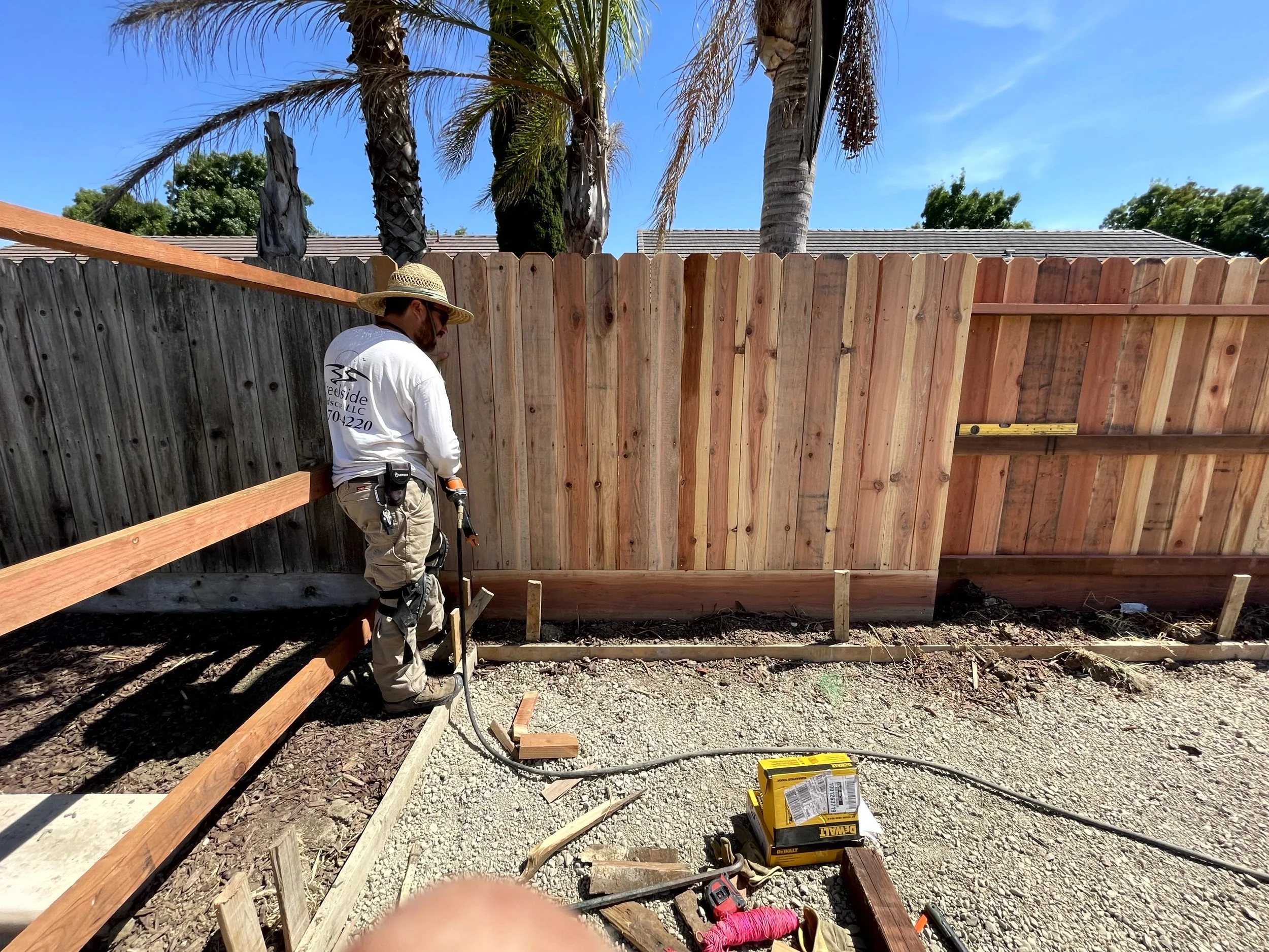 A construction worker installing a wooden fence in a backyard, wearing a hat, standing next to a finished black fence and partially finished natural wood fence, with tools on the ground and a bright sunny sky.