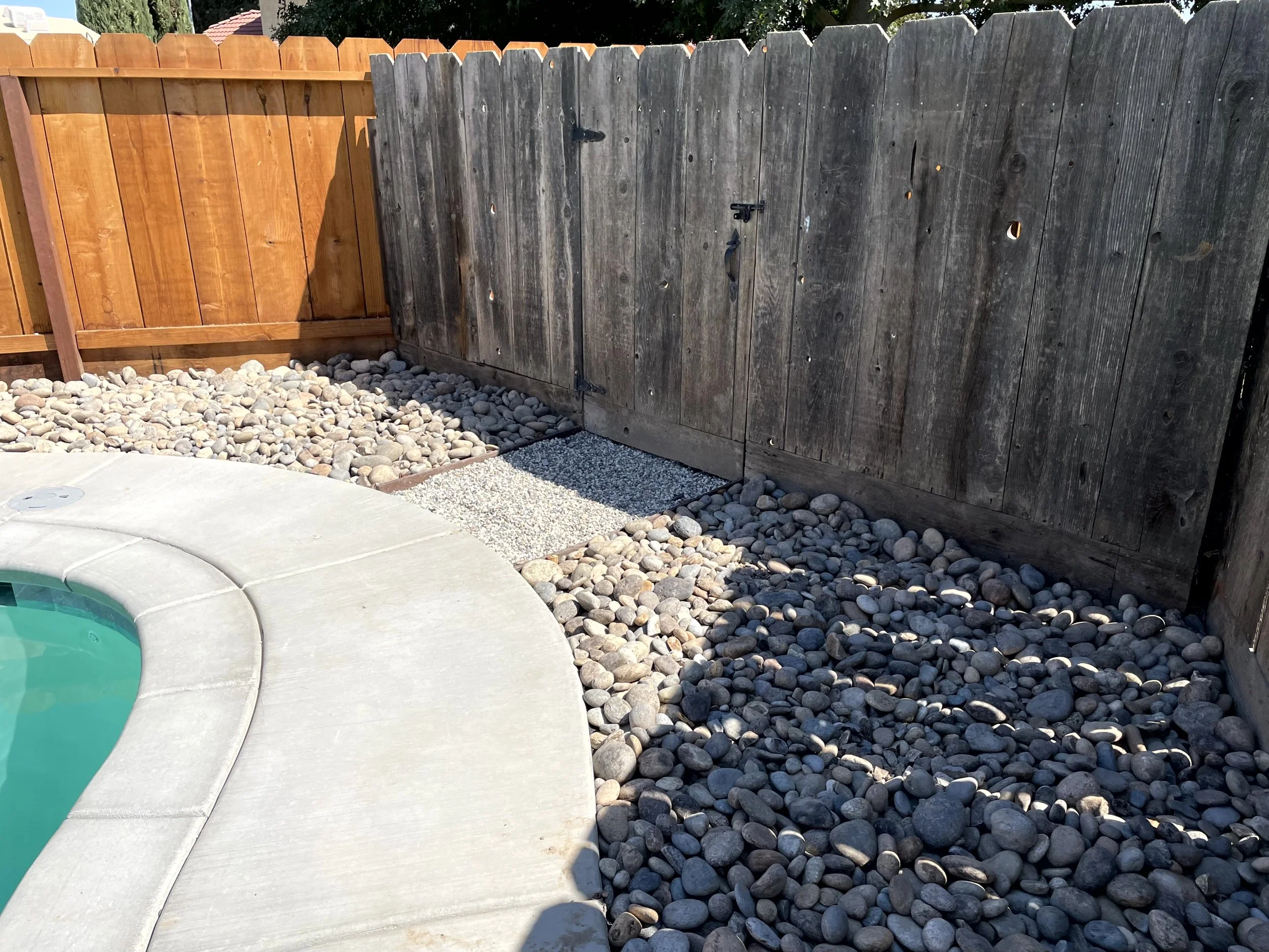 Corner of a backyard with a swimming pool, wooden and weathered fence, and decorative rocks.