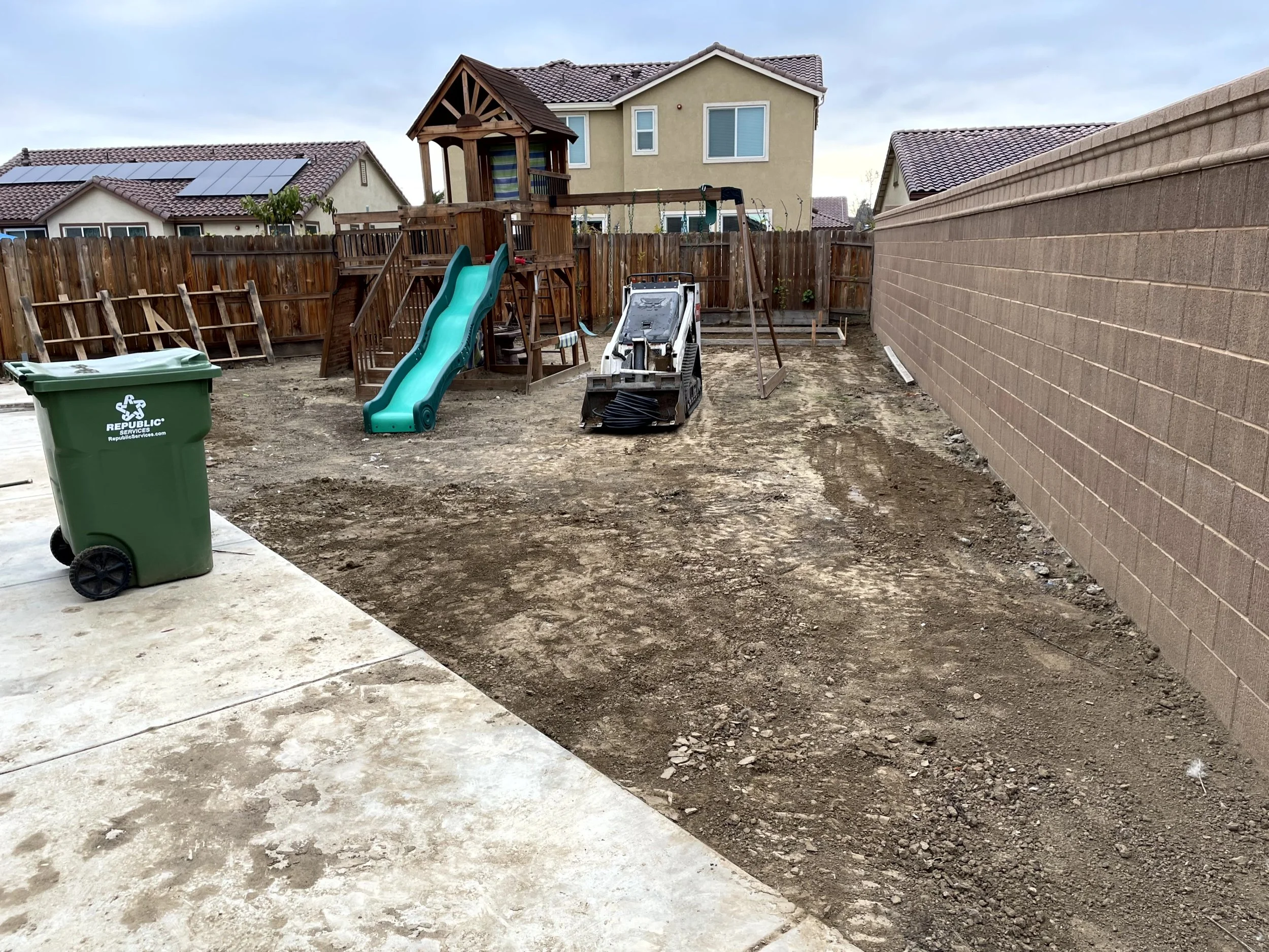 Backyard with a children's wooden playset that has a slide, with some construction equipment and dirt patches, next to a brick wall and a wooden fence, with neighboring houses in the background.