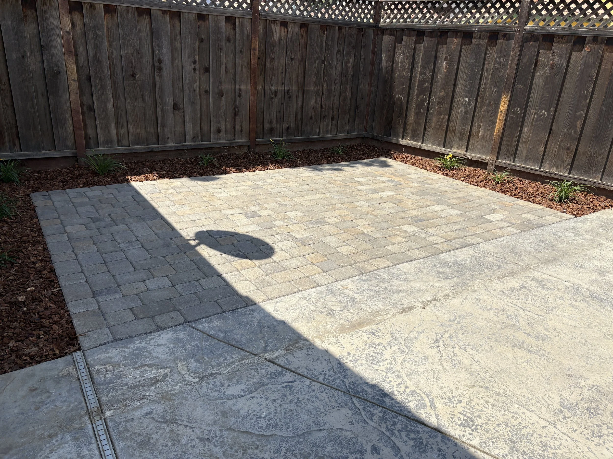 A backyard patio with a section of stone pavers, bordered by a flower bed with small plants, and a wooden fence in the background.
