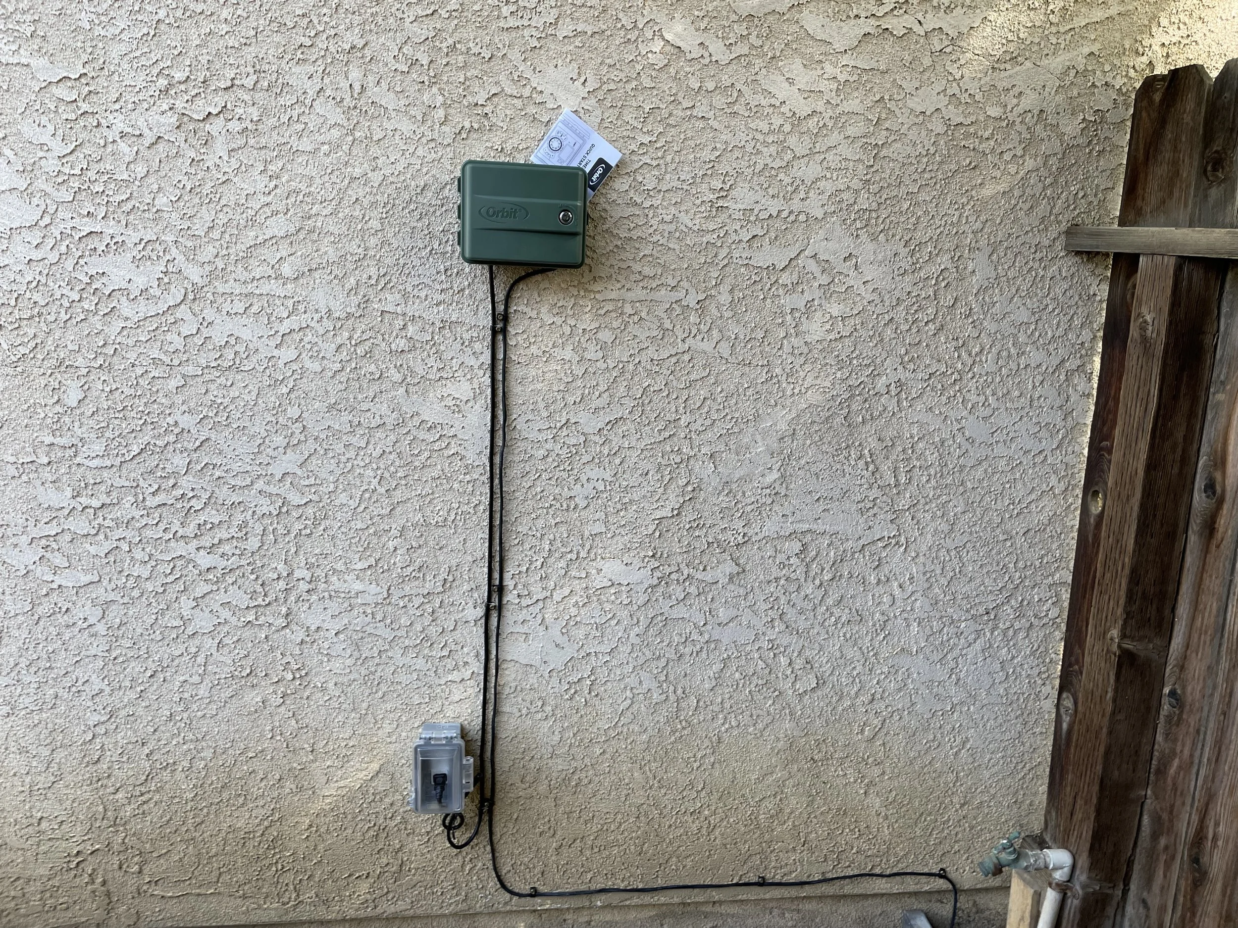 An outdoor beige textured stucco wall with a green electric meter, a black cable, a gray electrical box, and a water faucet with pipe on a wooden fence.