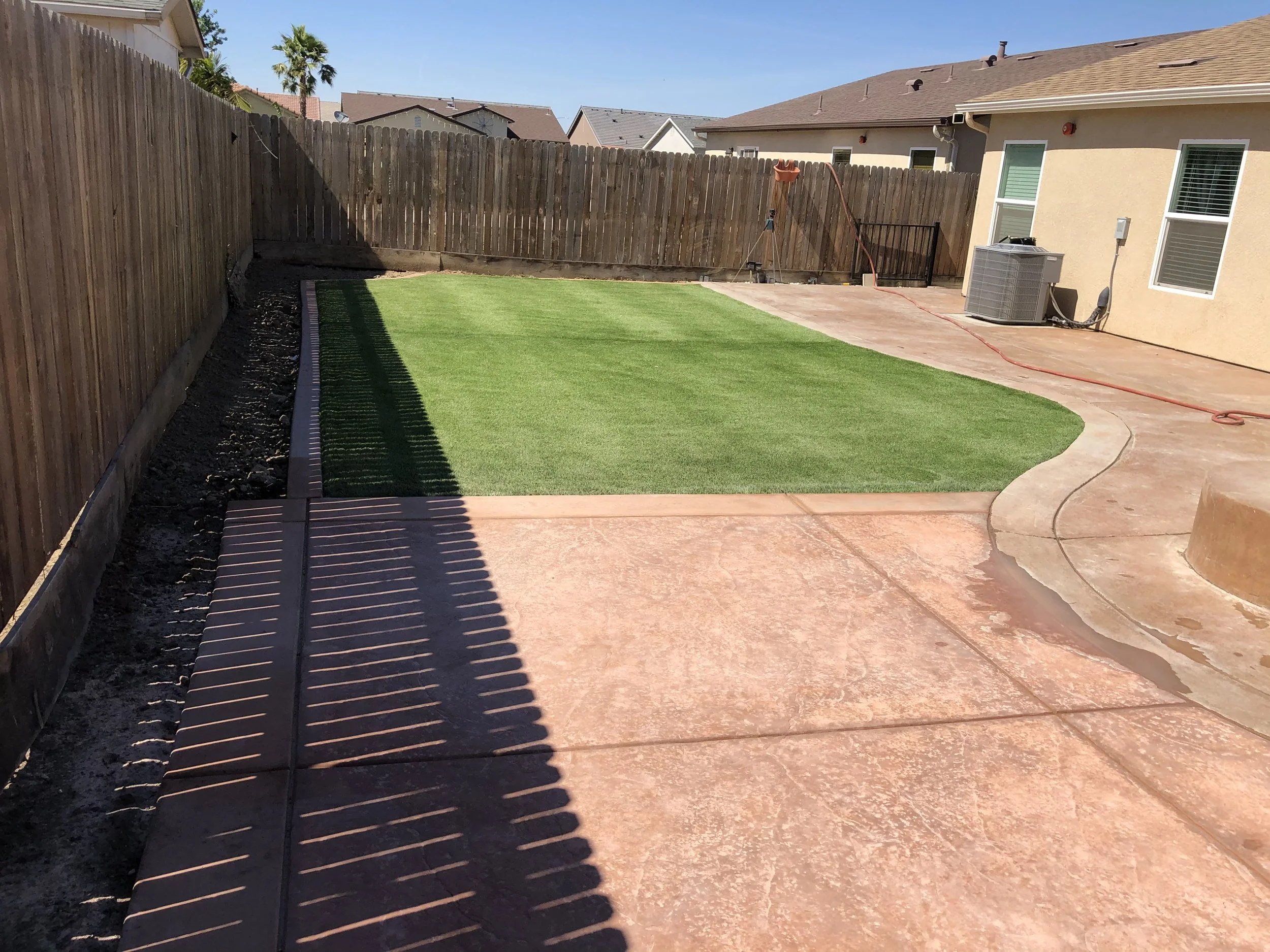 Backyard with a small green grass patch, a wooden fence, a concrete patio, and a house with windows and an air conditioning unit.