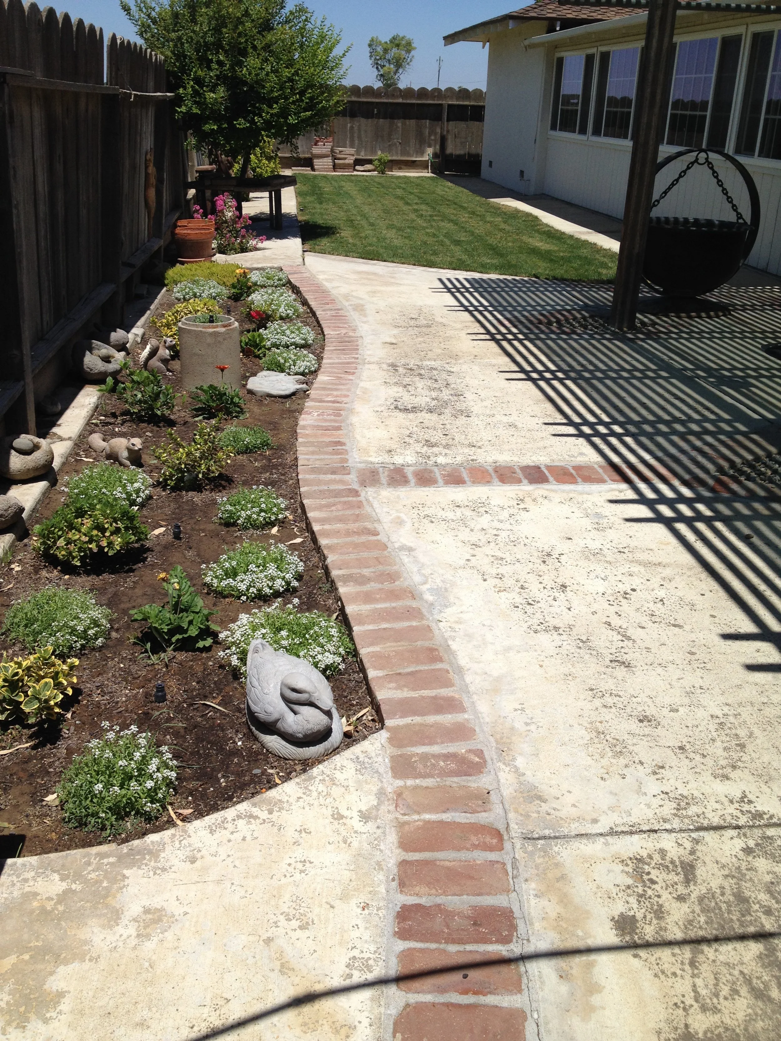Backyard with a curved brick border, garden bed with flowers and garden ornaments, a lawn, and a house with a shaded porch and swing.