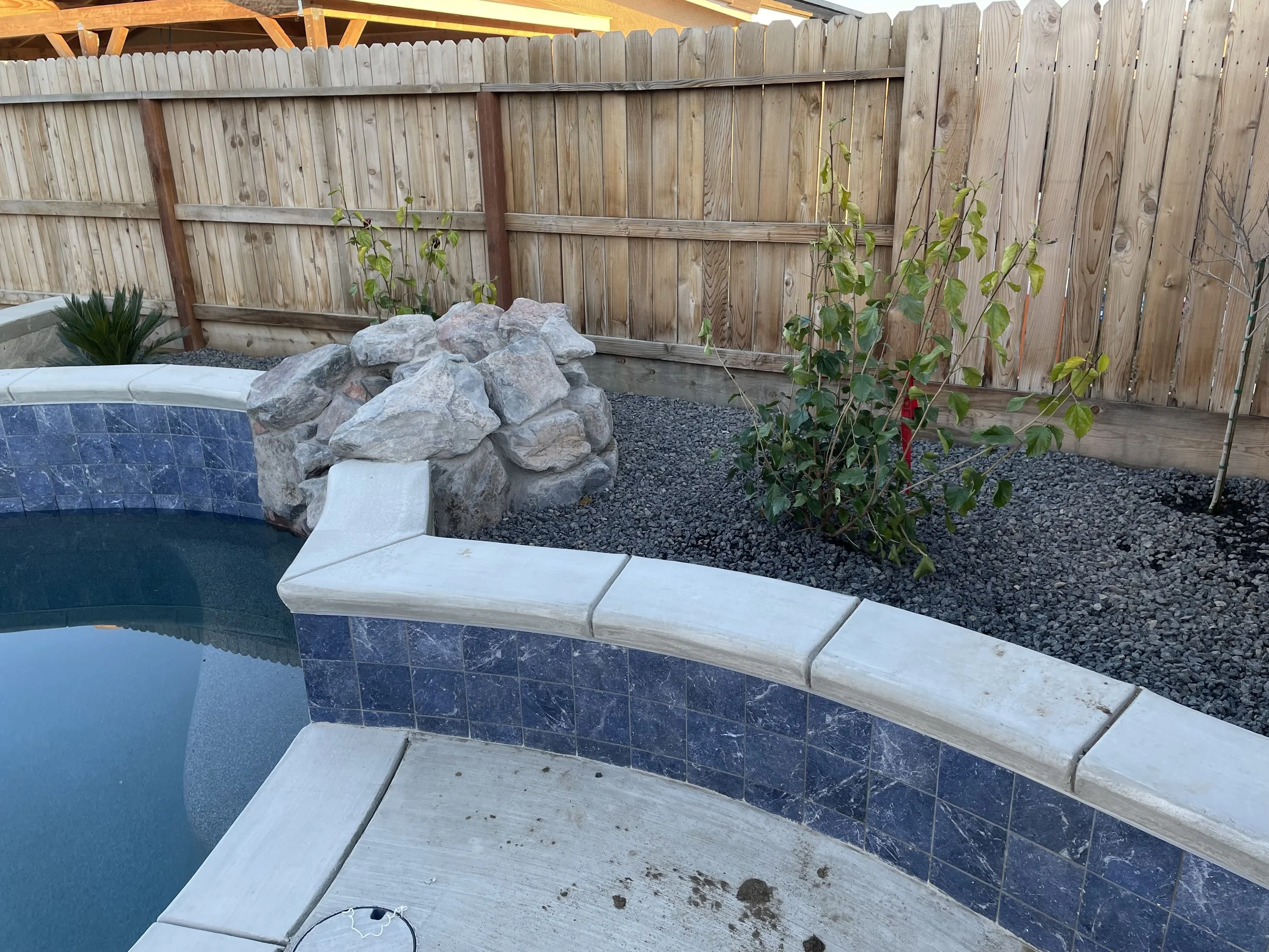Backyard with a swimming pool, rock waterfall feature, and a small plant bush with a wooden fence in the background.