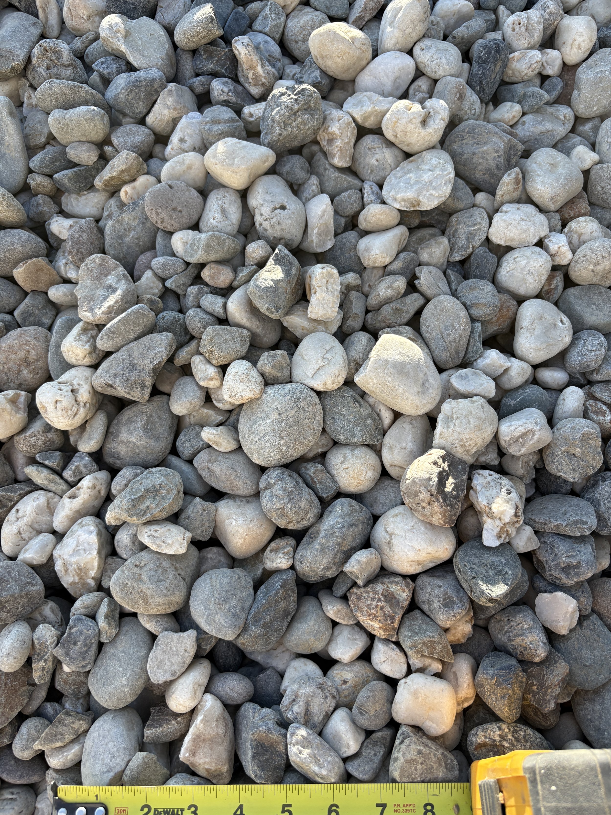 Close-up view of various small, smooth gray and beige stones and pebbles on the ground, with a yellow measuring tape at the bottom.