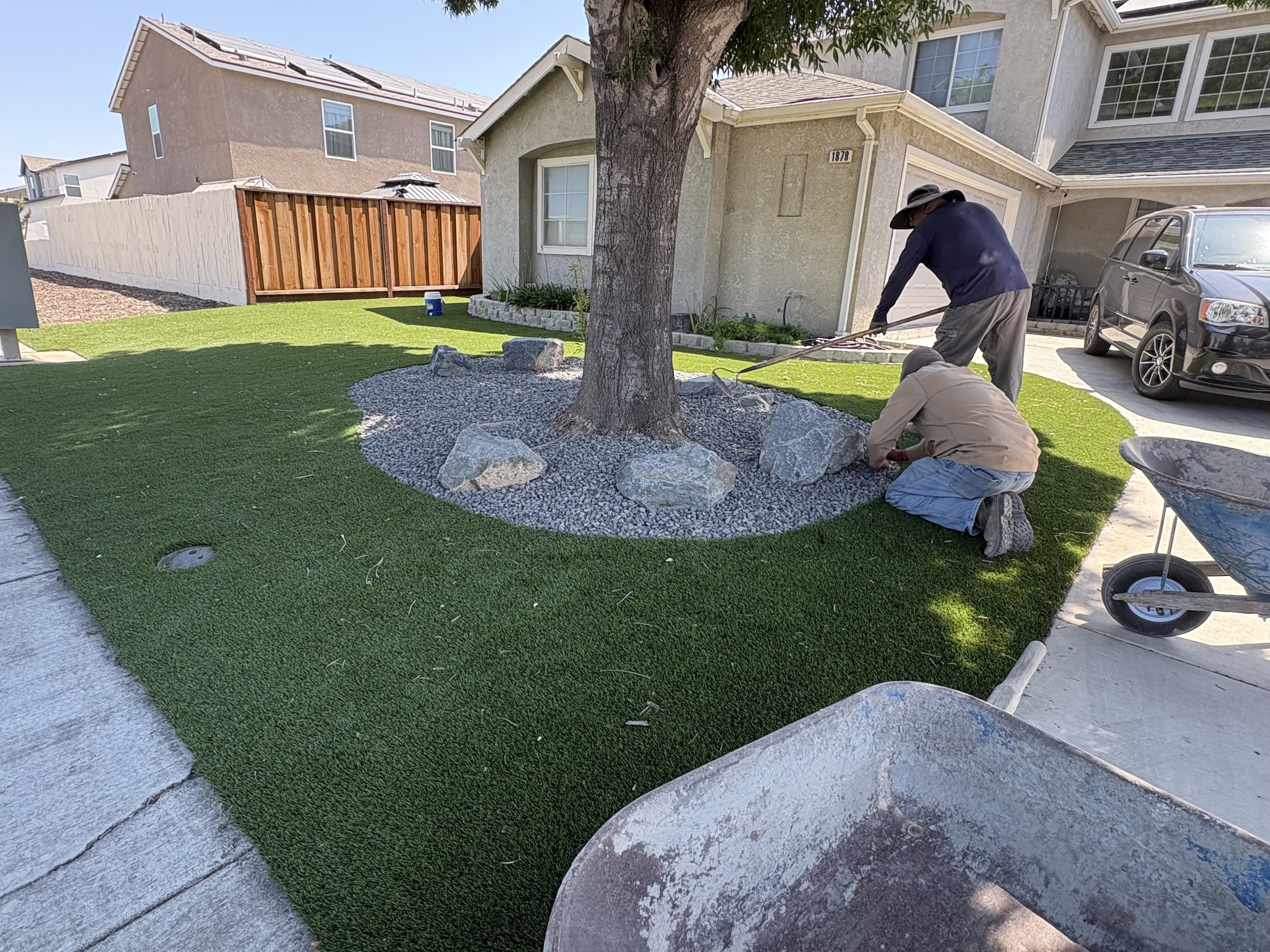 Two workers planting and arranging rocks around a tree in a front yard with lush green grass, a house in the background, and a wheelbarrow nearby.