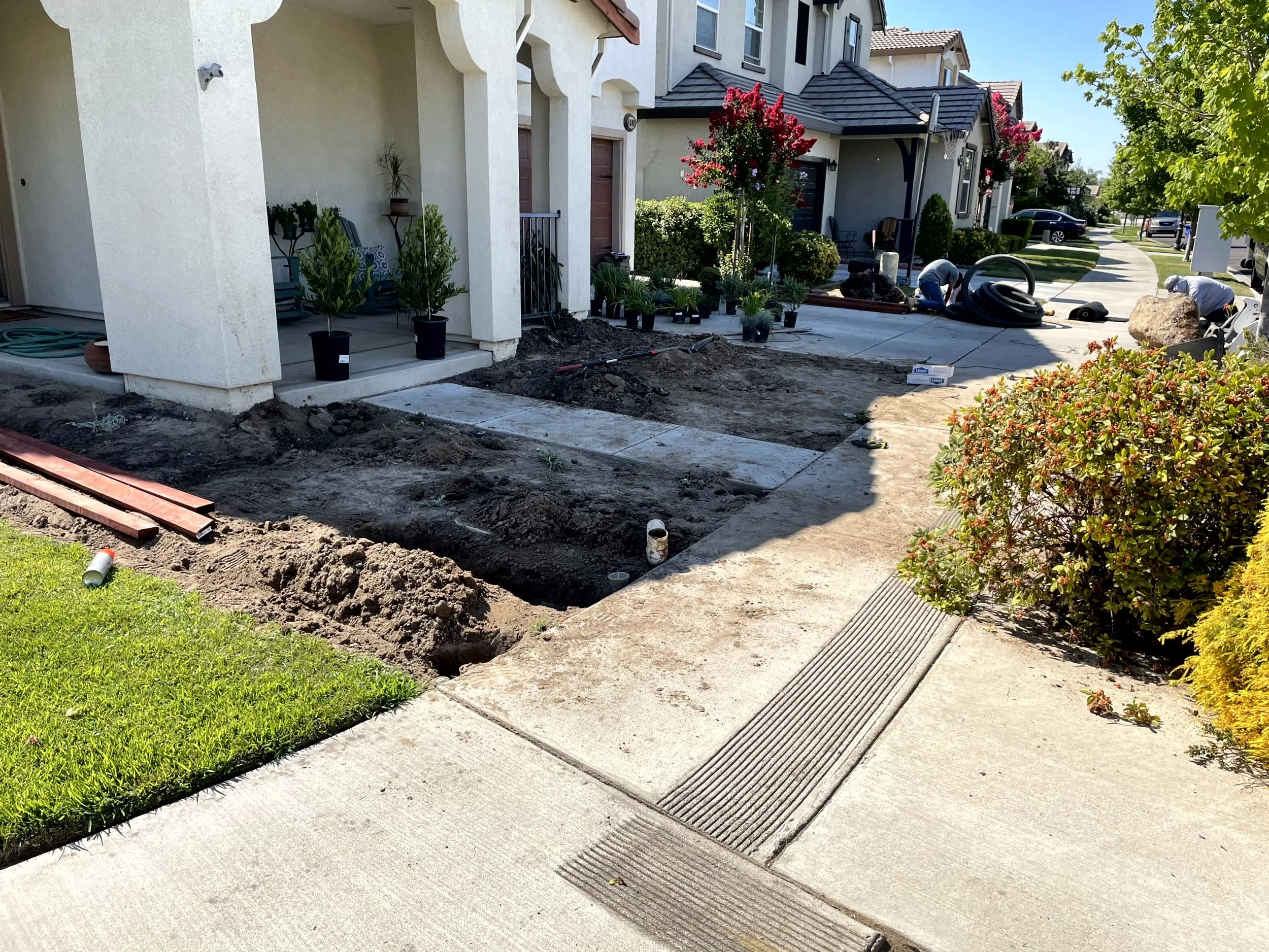 Front yard of a residential neighborhood under construction, with dirt, tools, plants, and a partially dug trench.