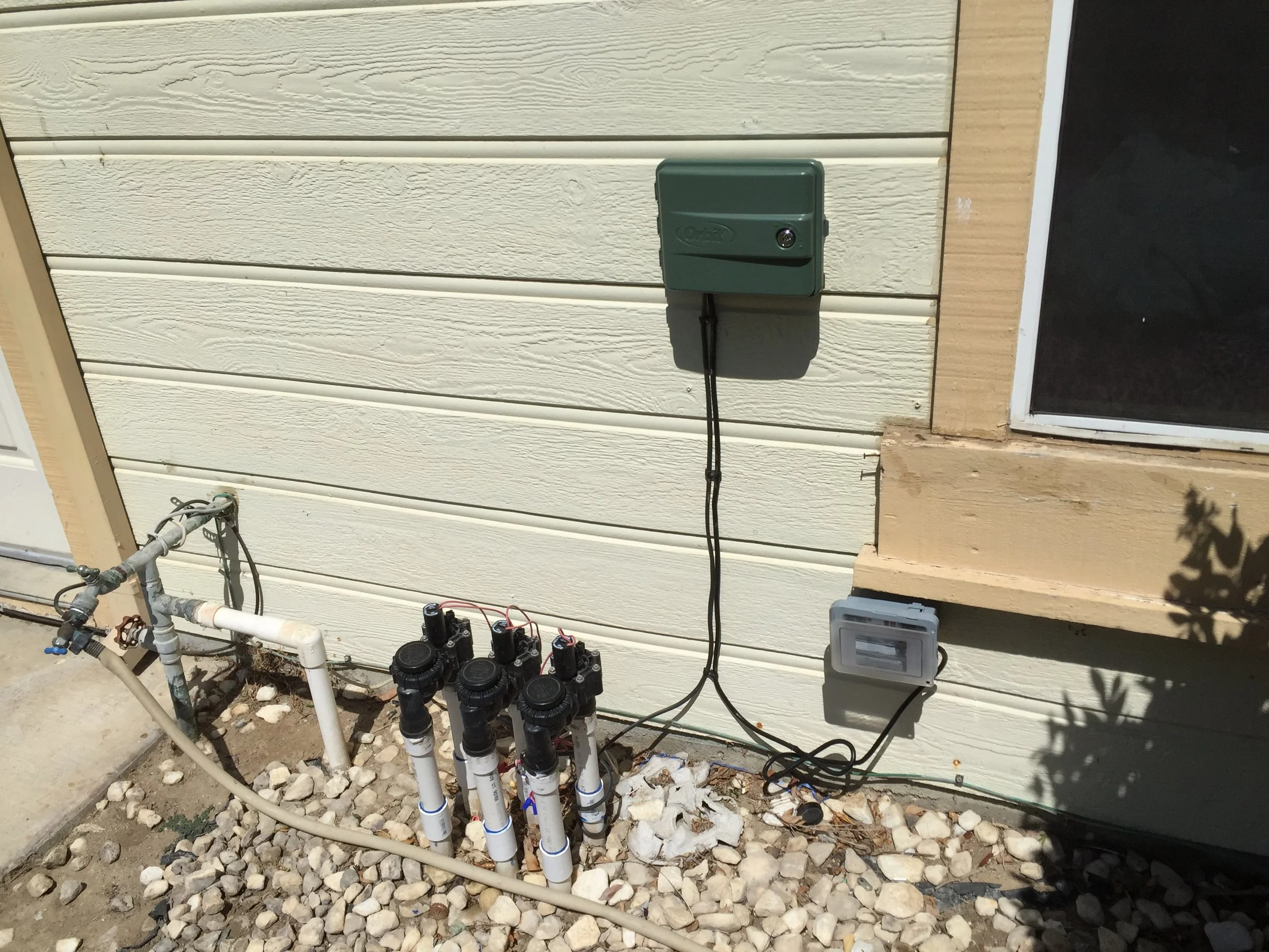 Outdoor view of a house with plumbing and electrical components, including a green electrical box, multiple black and white pipes, and various wire connections on a beige exterior wall with rocks and dirt below.