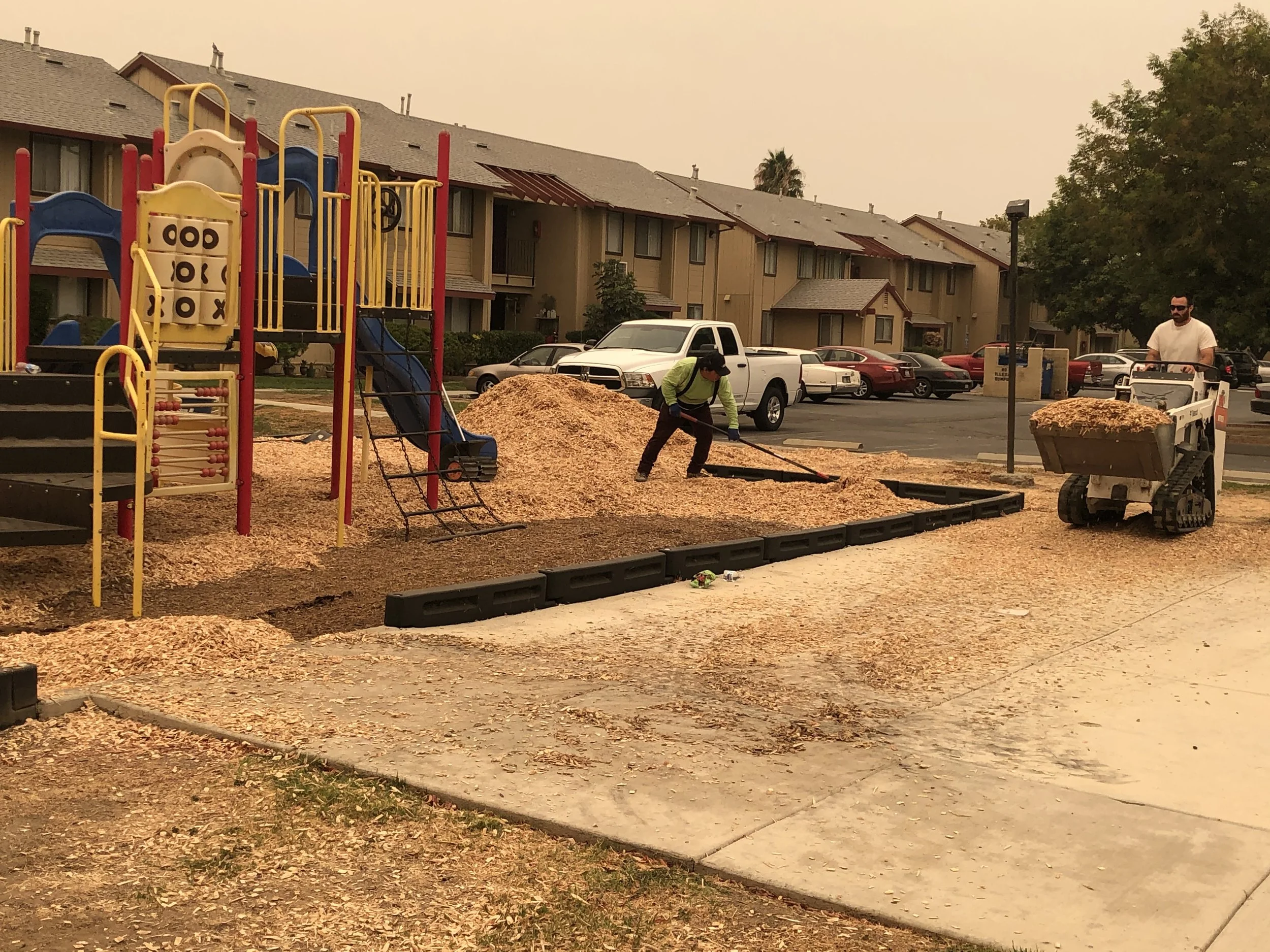 Workers installing new playground surface with wood chips in a residential park, with playground equipment and parked cars in the background.