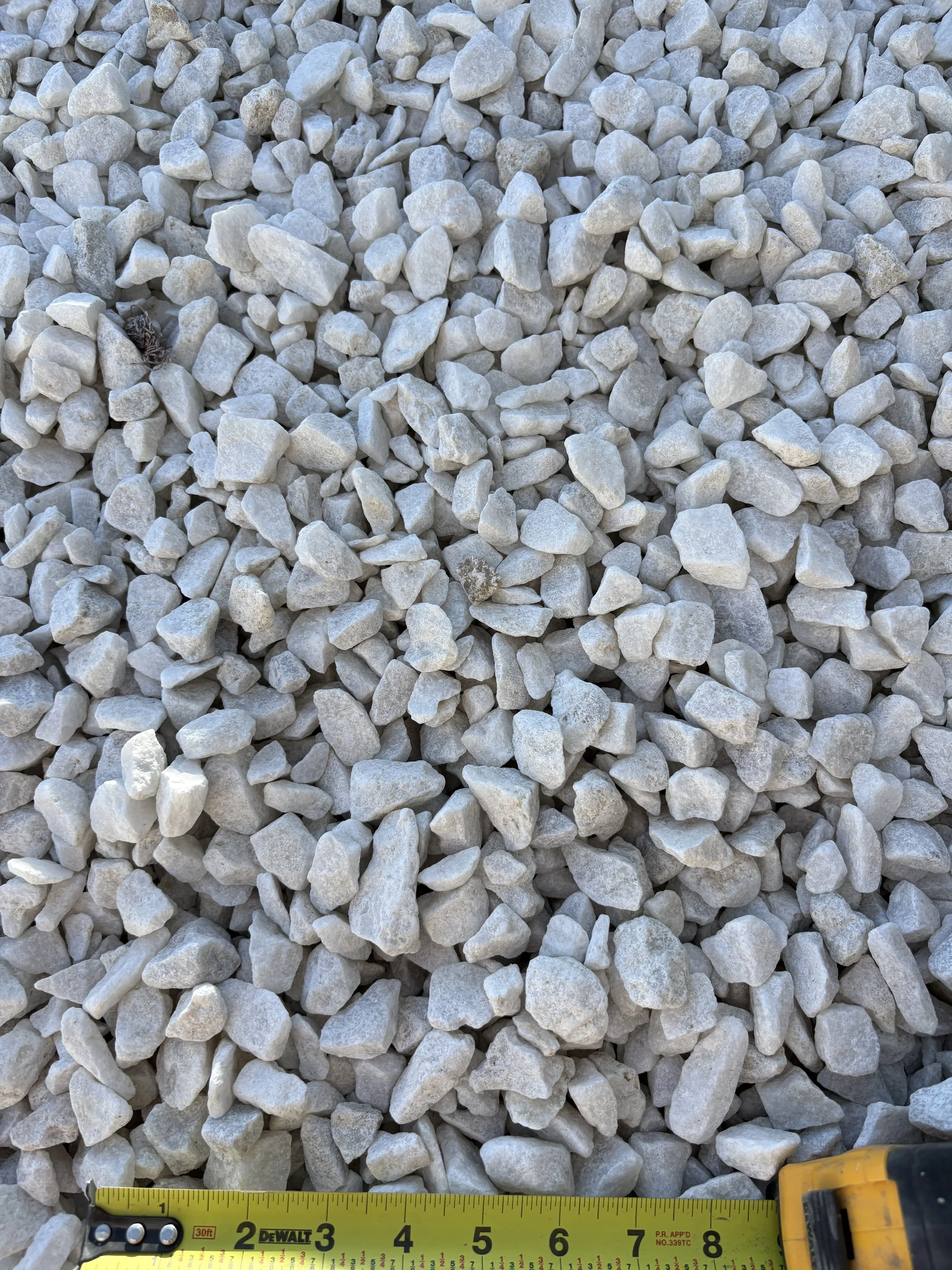 Close-up of small gray and white gravel stones, with a yellow measuring tape at the bottom showing measurements in inches.