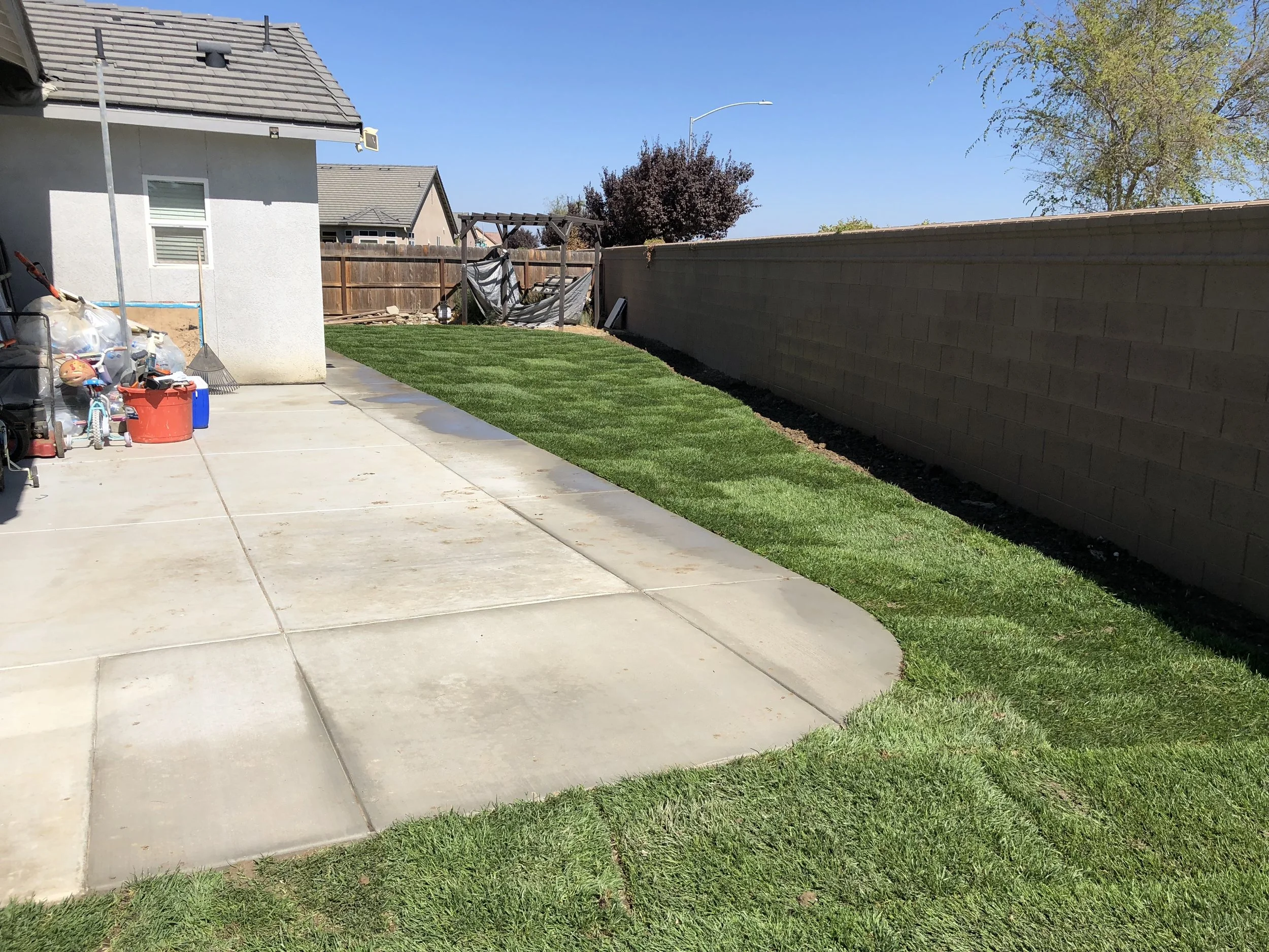 Backyard with a concrete patio, a grassy lawn, a wooden fence, and a privacy wall; some toys and gardening supplies near the house.