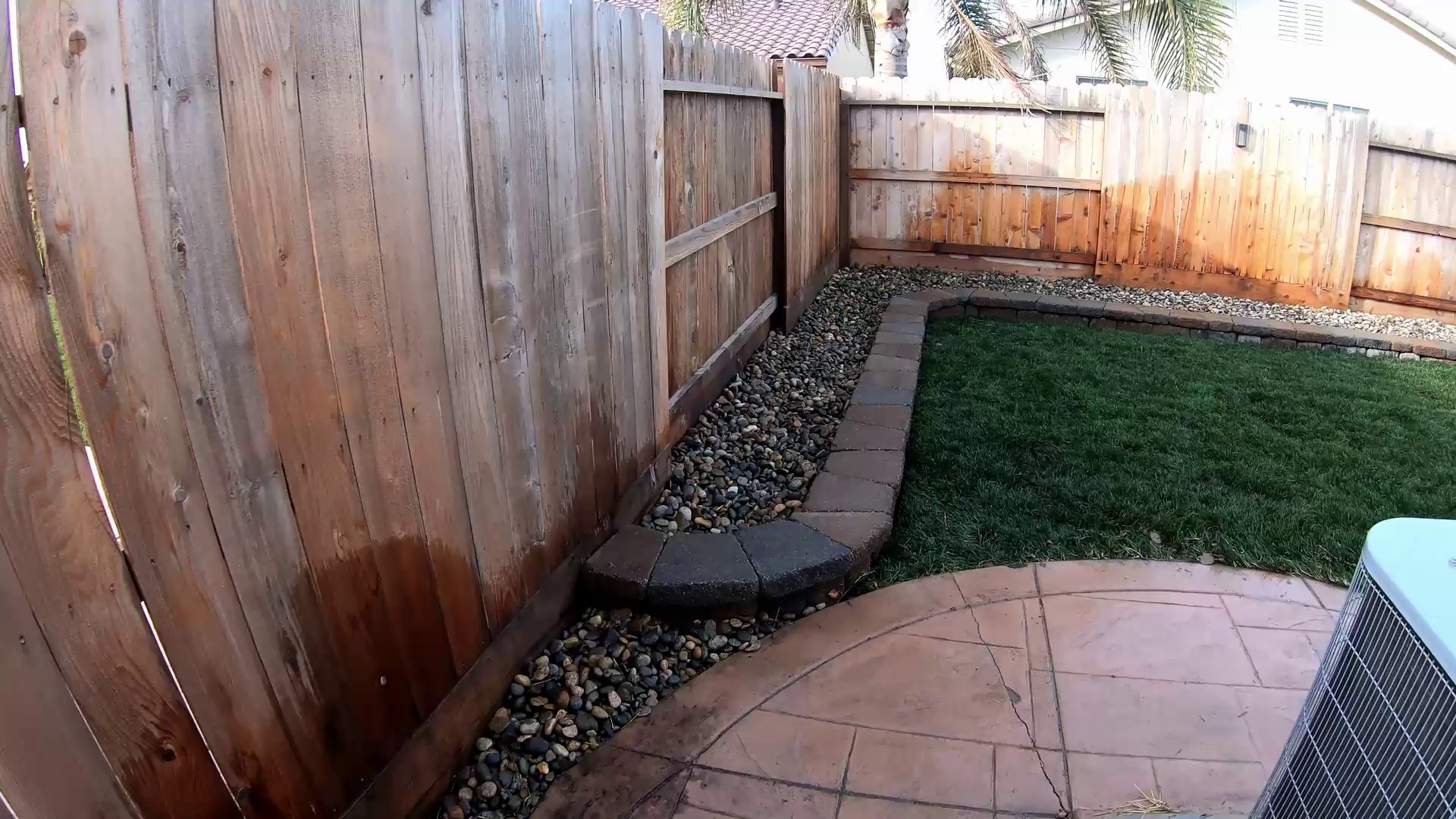 A backyard with a wooden fence, a small patch of green grass, and a curved edge with pavers and rocks, seen from a surveillance camera.
