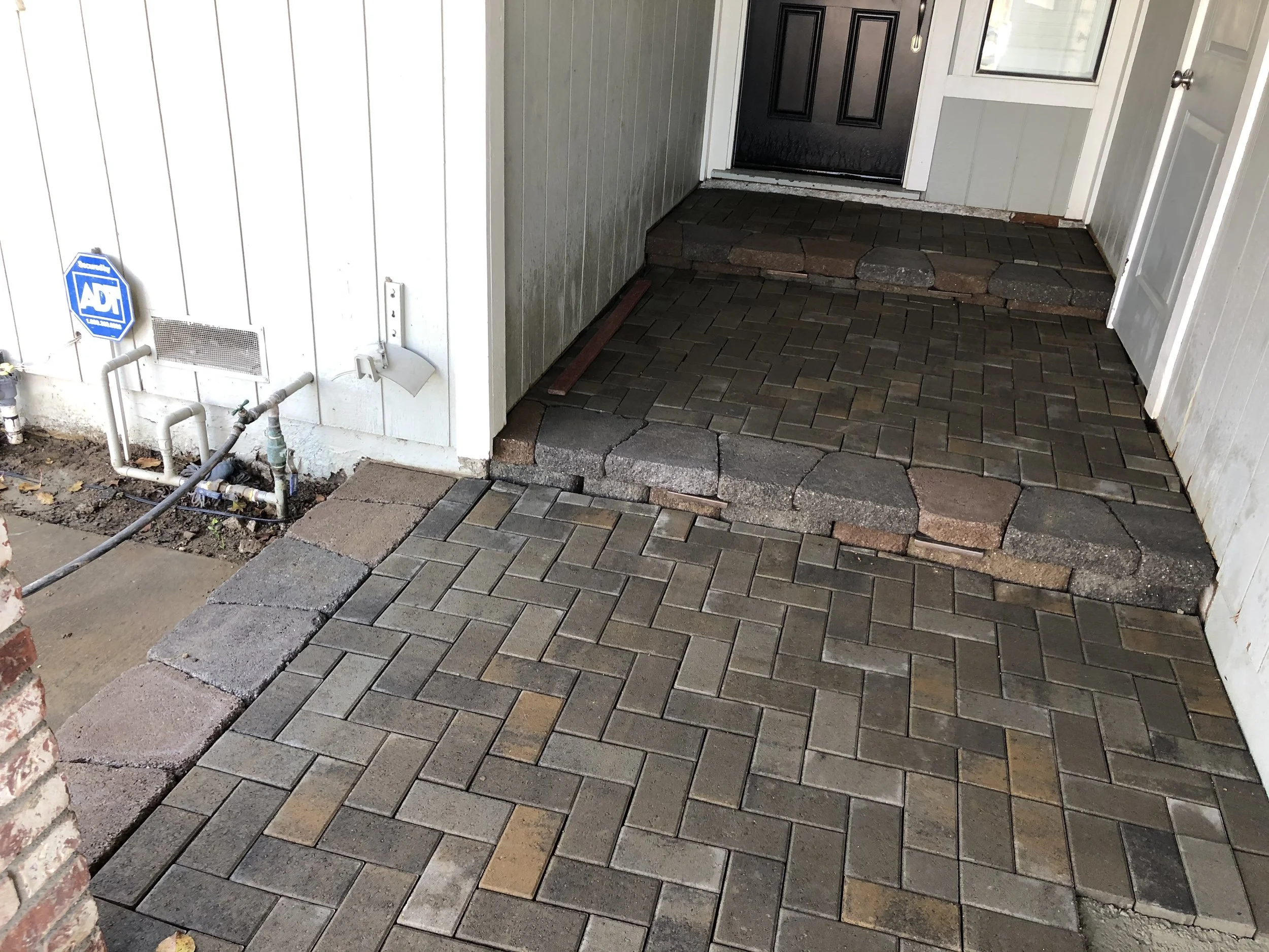 Newly installed paver walkway with border around the front door, featuring gray, tan, and reddish bricks, leading to a black front door.