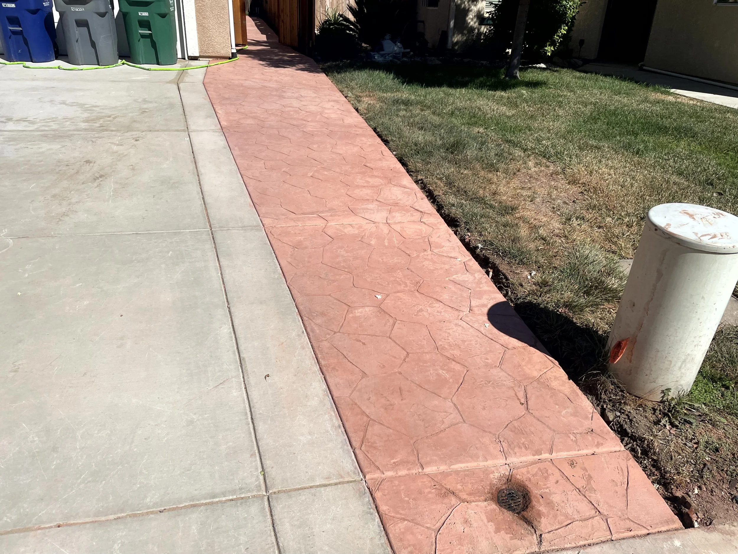 Sidewalk with a concrete section and a red stamped concrete section, next to a grassy area with a white bollard and a storm drain.