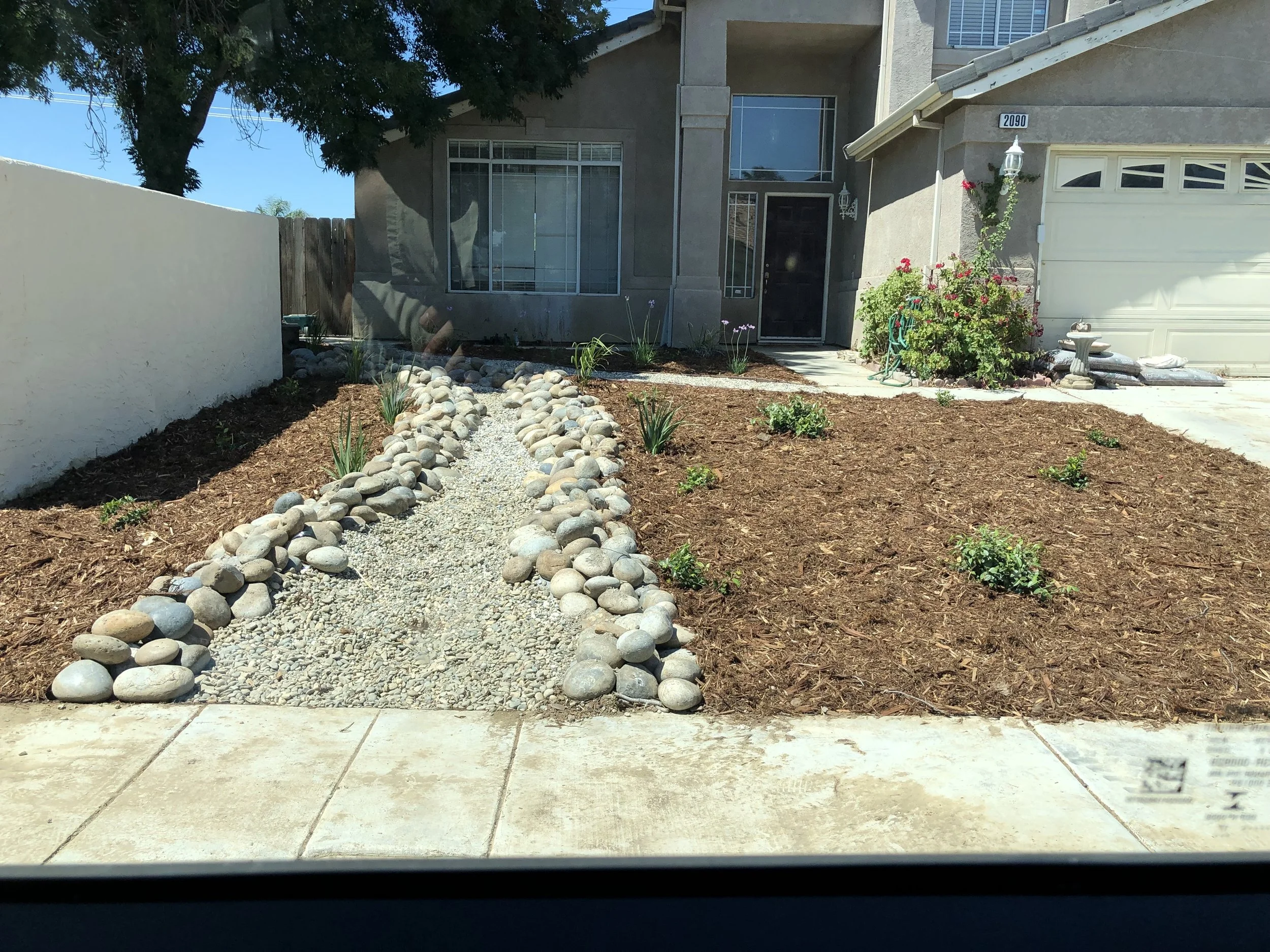 Front yard landscape with a dry creek bed of small gravel and large rounded rocks, young plants, and a patch of brown mulch in front of a house with large windows and a garage.