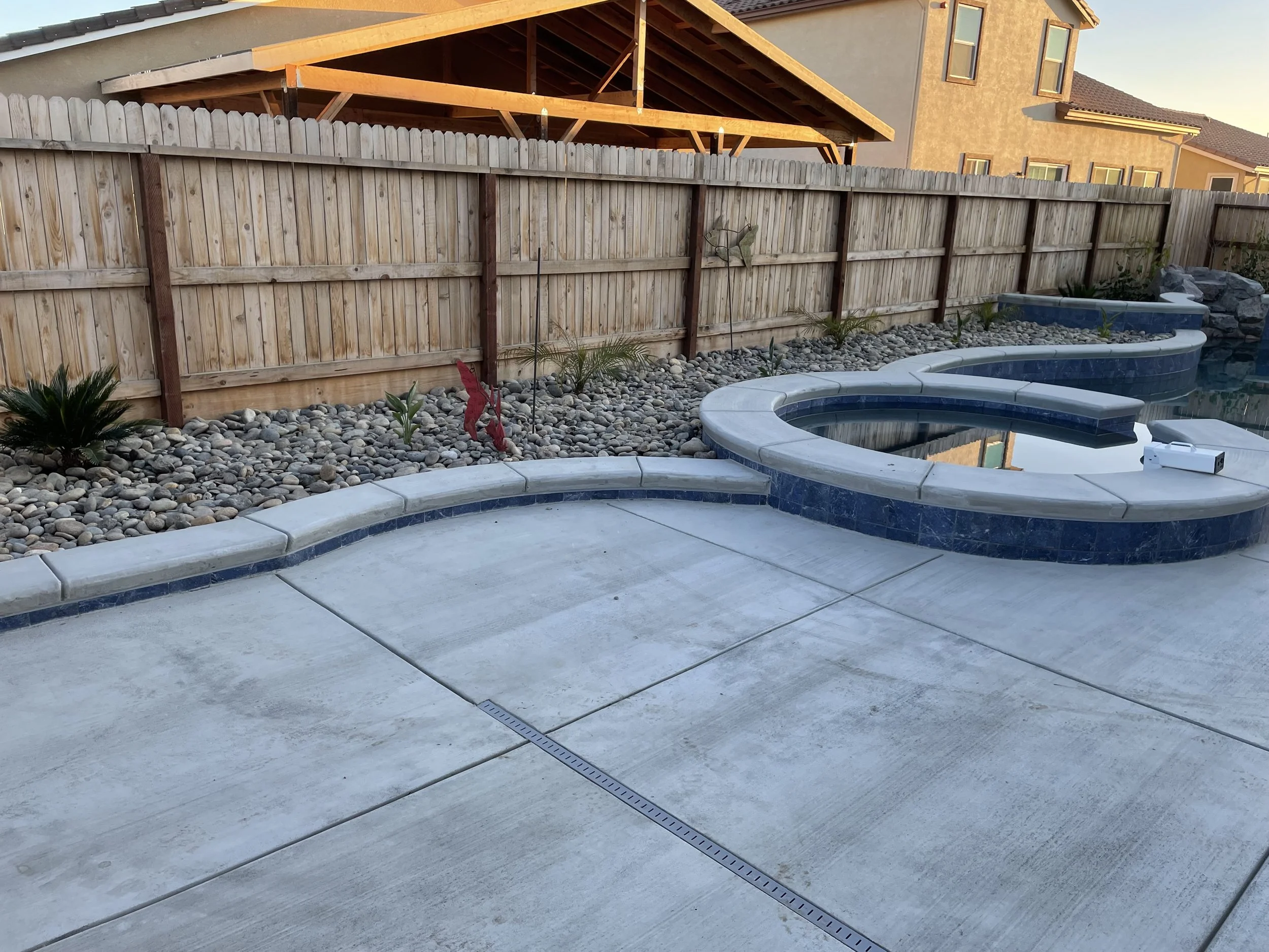 Backyard with a swimming pool, a wooden fence, and some small plants. The pool has a curvy shape with a small fountain or feature in the middle. The area looks tidy and well-maintained.