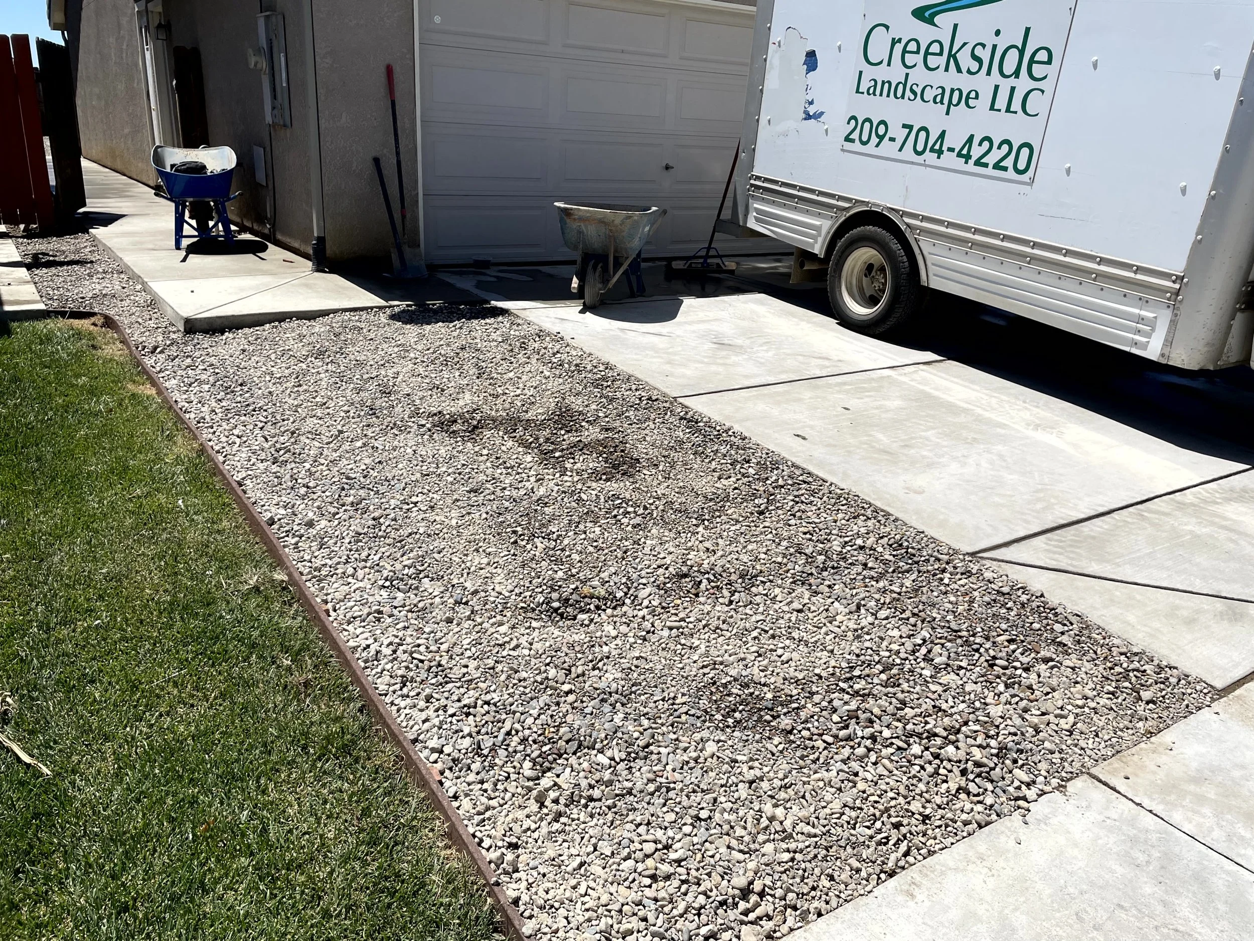 A concrete driveway under construction with gravel and stone border. Nearby are construction tools, a wheelbarrow, and a moving truck with 'Creekside Landscape LLC' logo.