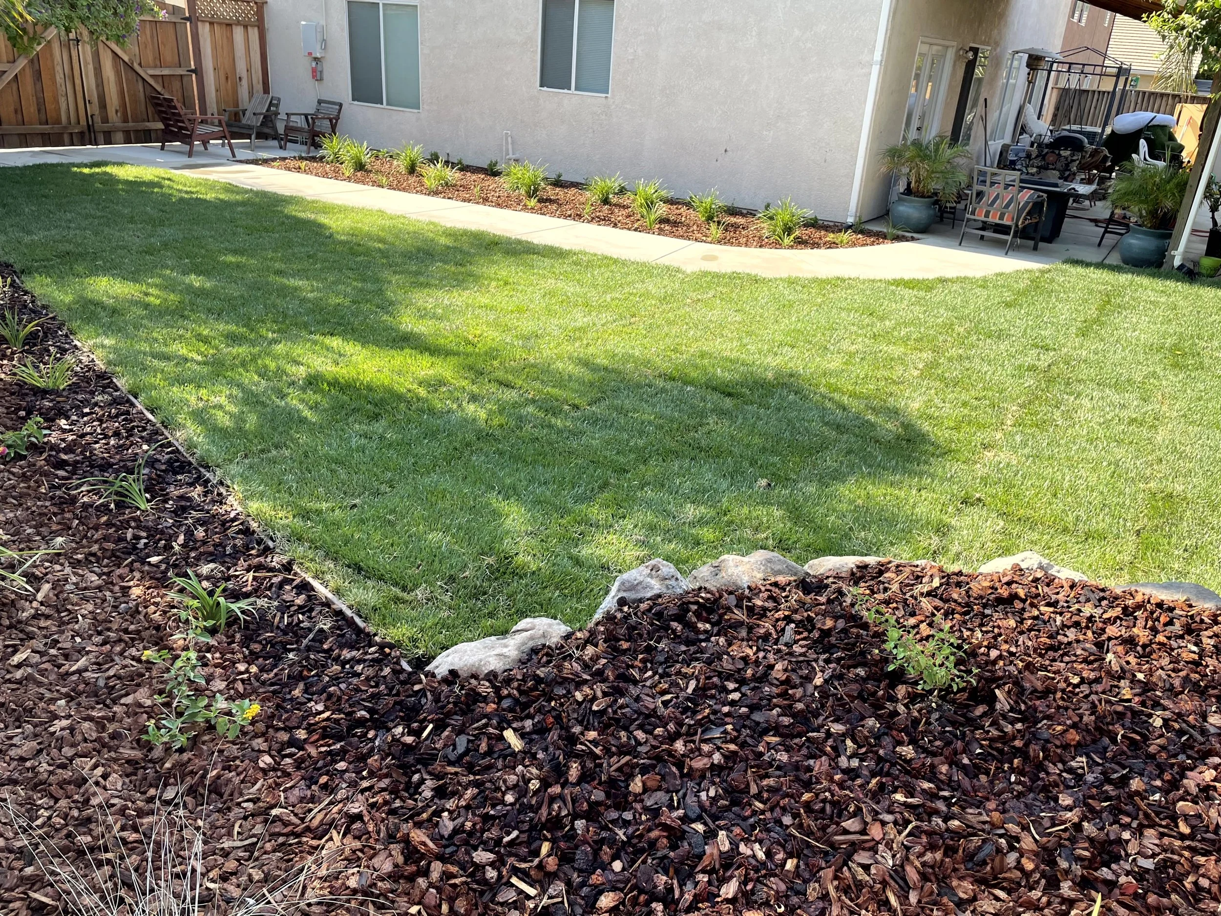 Backyard with a well-maintained green lawn, garden beds with mulch, small plants, and a concrete patio with outdoor furniture, including chairs and potted plants.