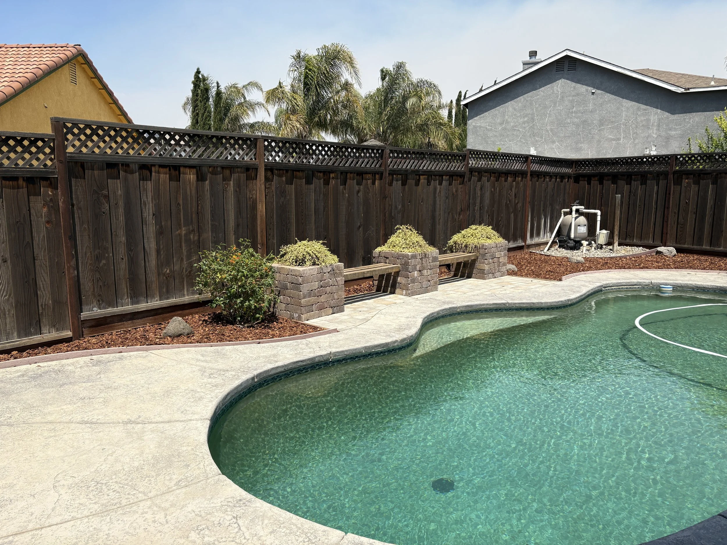 Backyard with a swimming pool, wooden fence, potted plants, and a pool filter system in the corner.