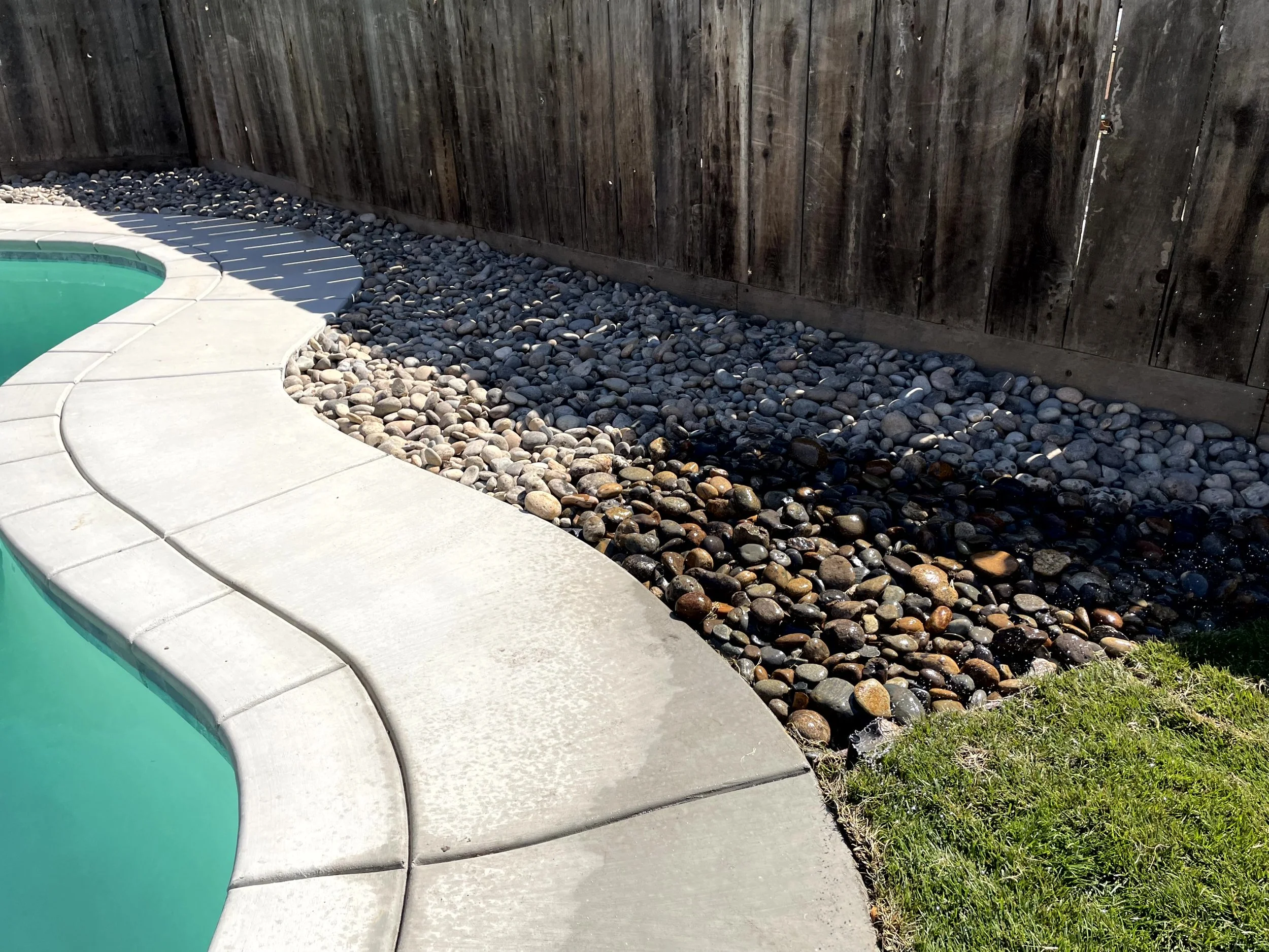 Curved concrete pool deck next to a small patch of green grass, with a rock border and a wooden fence in the background.