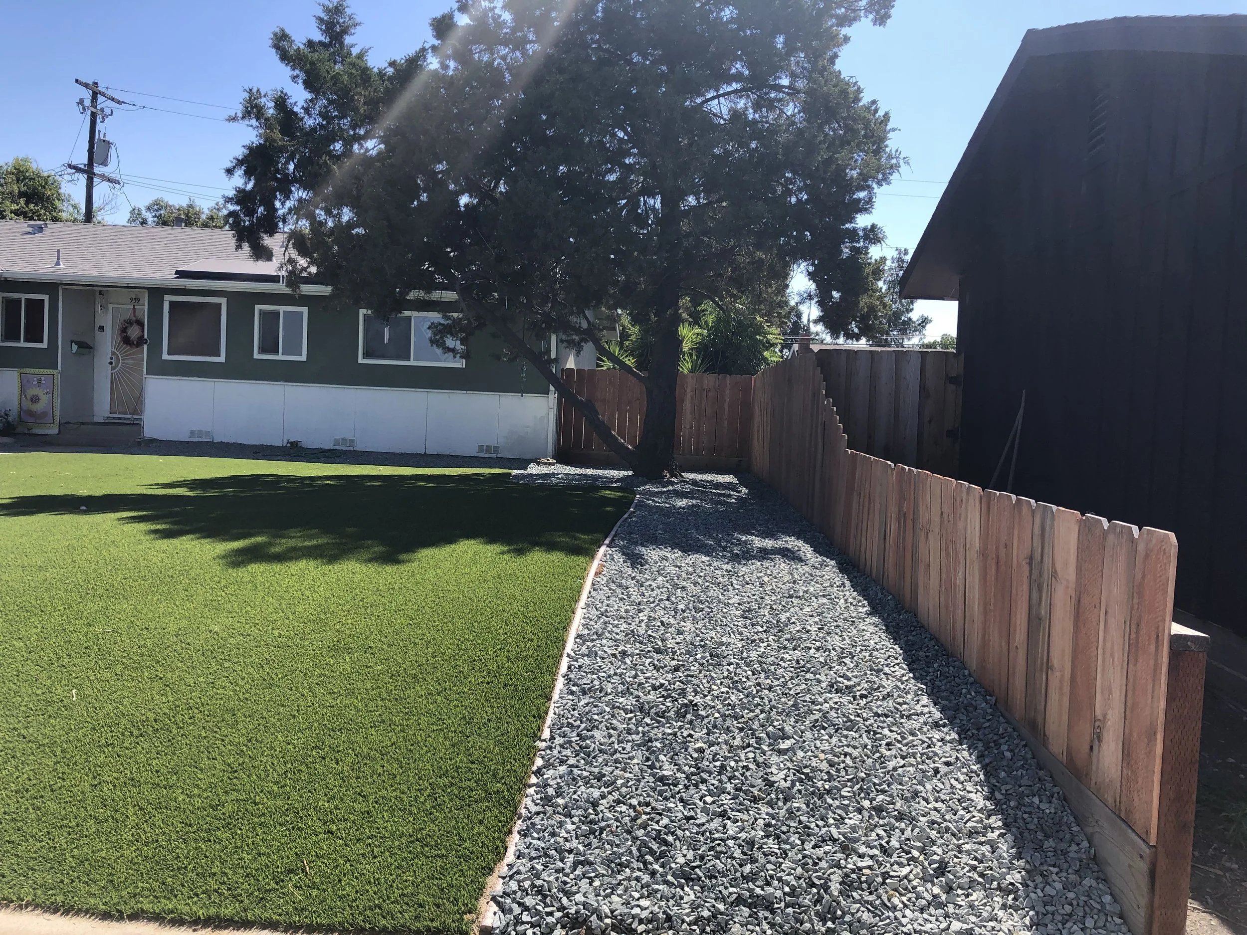 A backyard with a well-maintained green lawn, a gravel pathway, a large tree, and a wooden fence separating neighboring properties under a clear blue sky.