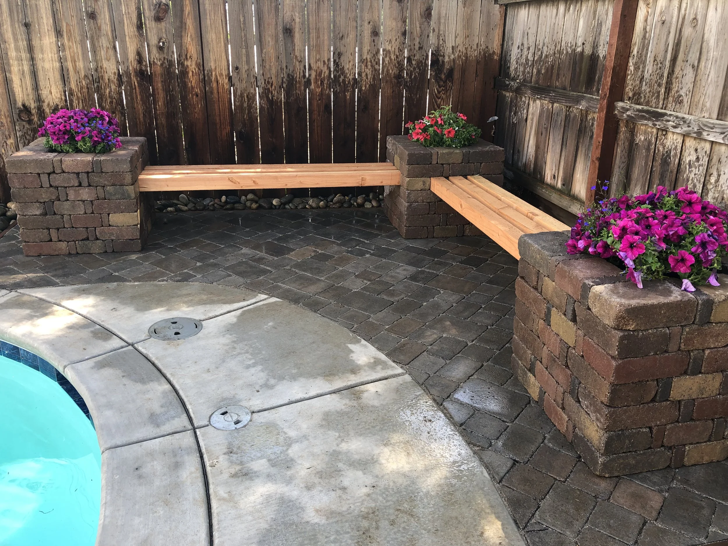 A backyard corner with two brick planters holding pink and purple flowers, and wooden benches between them, alongside a wooden fence and a pool edge.