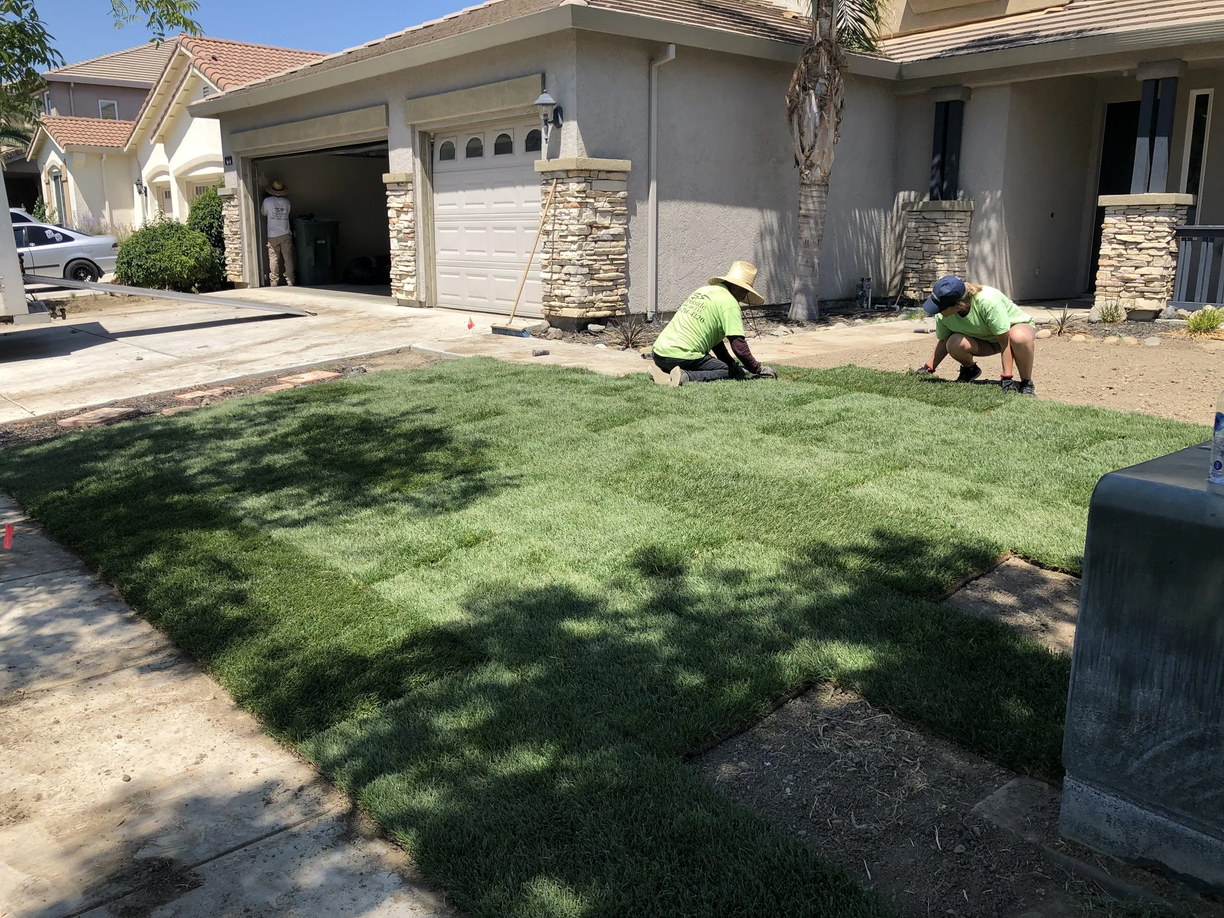 Two workers planting grass in a residential front yard while another person stands near a garage in the background. The house has stone accents and a tree provides shade.