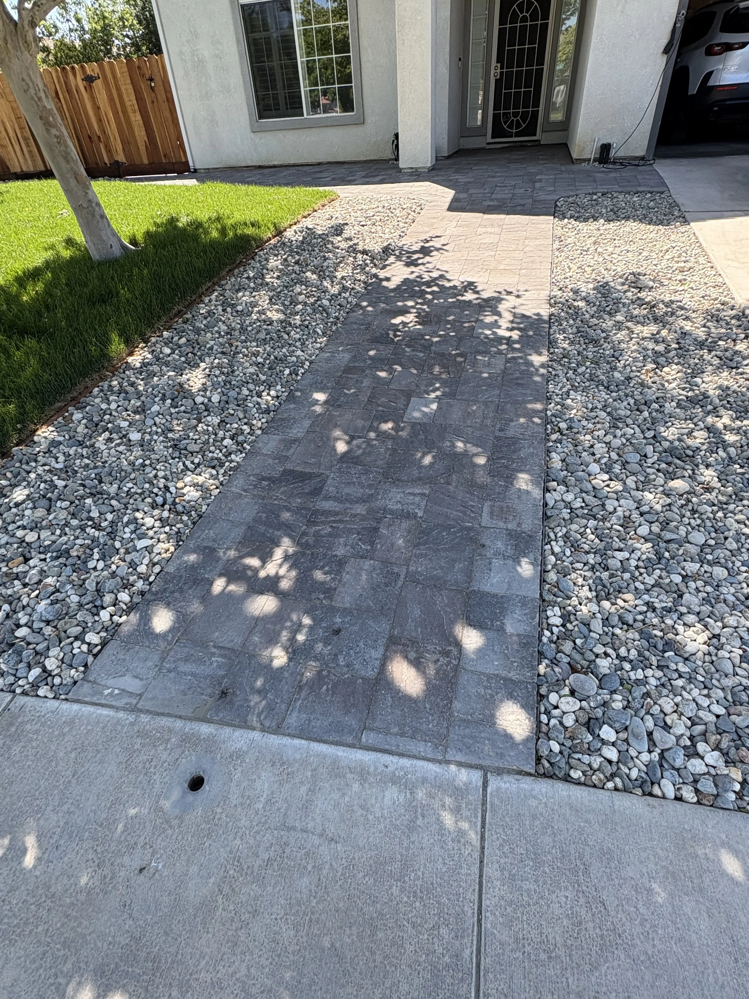 Residential front yard with a pathway leading to the house, surrounded by decorative rocks and a strip of grass with a tree, in front of a white house with a front door and a driveway with a car.