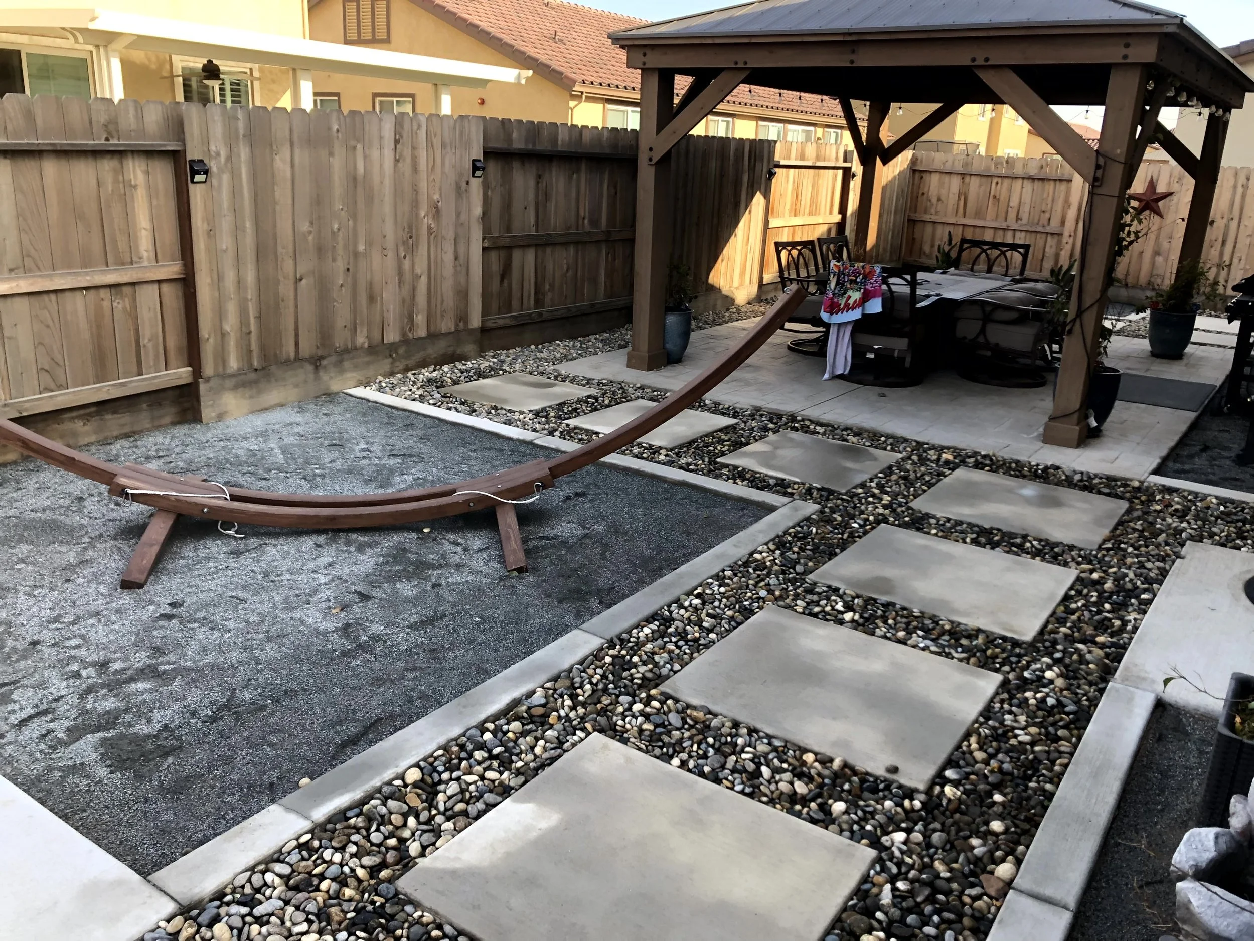 A backyard with a wooden fence, a sliding garden gate, a gravel area, concrete stepping stones set in pebble borders leading to a covered patio with outdoor furniture, and a wooden swing with a slide.