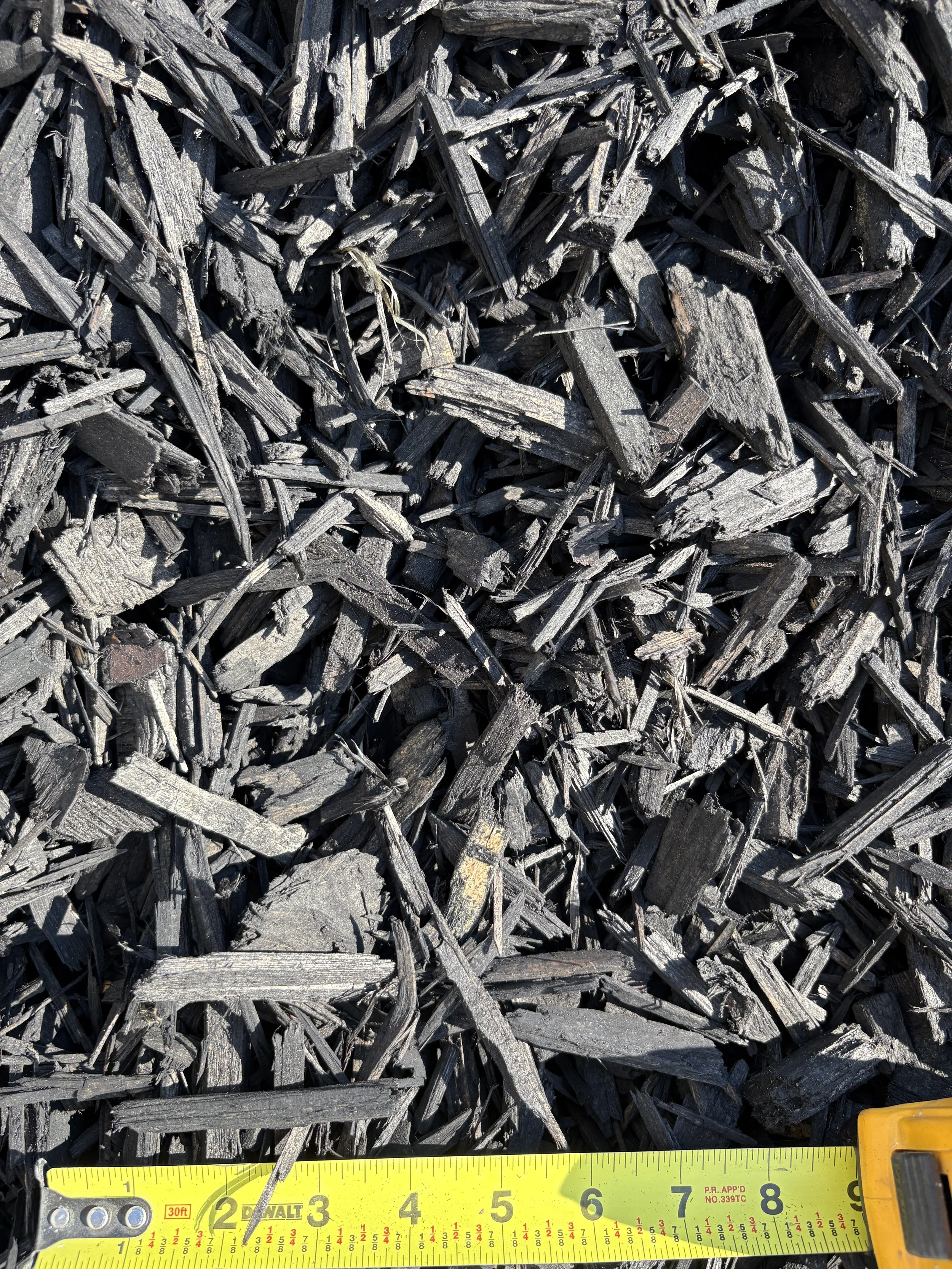 Close-up of black wood chips or mulch with a yellow measuring tape at the bottom showing measurement markings.