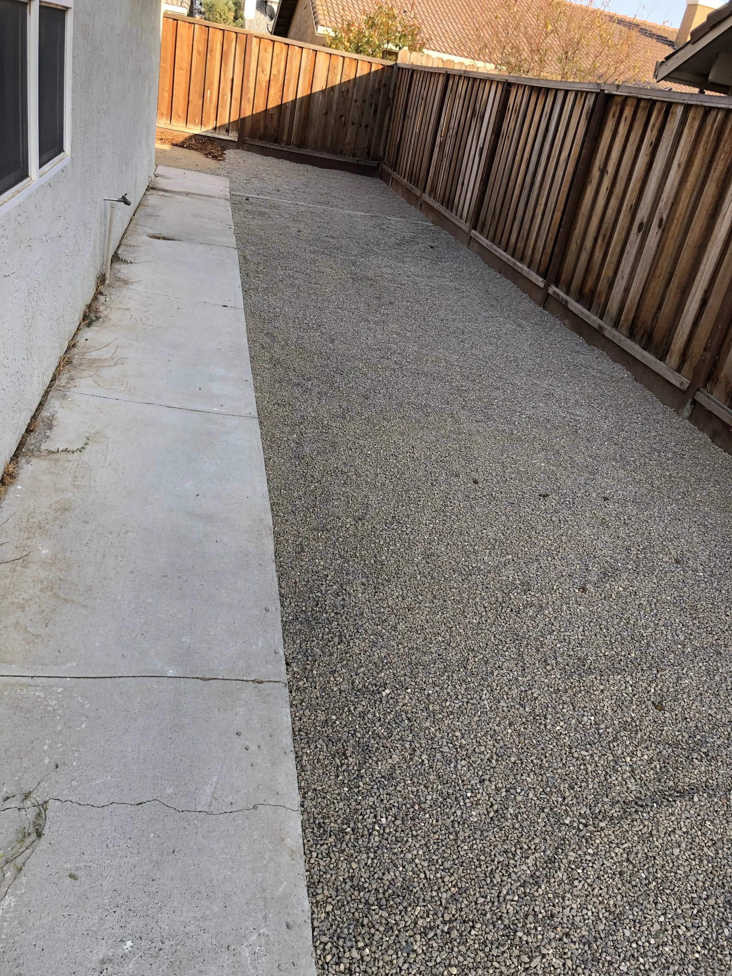 Backyard with gravel ground, concrete walkway, wooden fence, and side of house with window