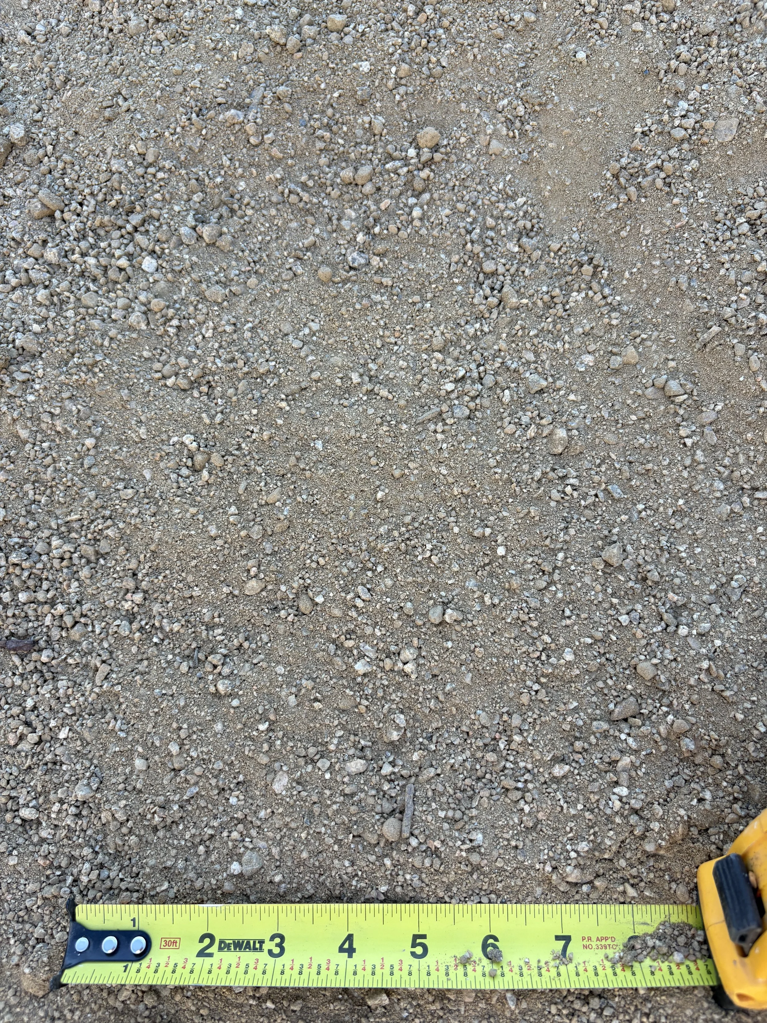 Close-up of sandy ground with small pebbles and rocks, a yellow measuring tape showing approximately 6 inches, and part of a yellow tool in the lower right corner.