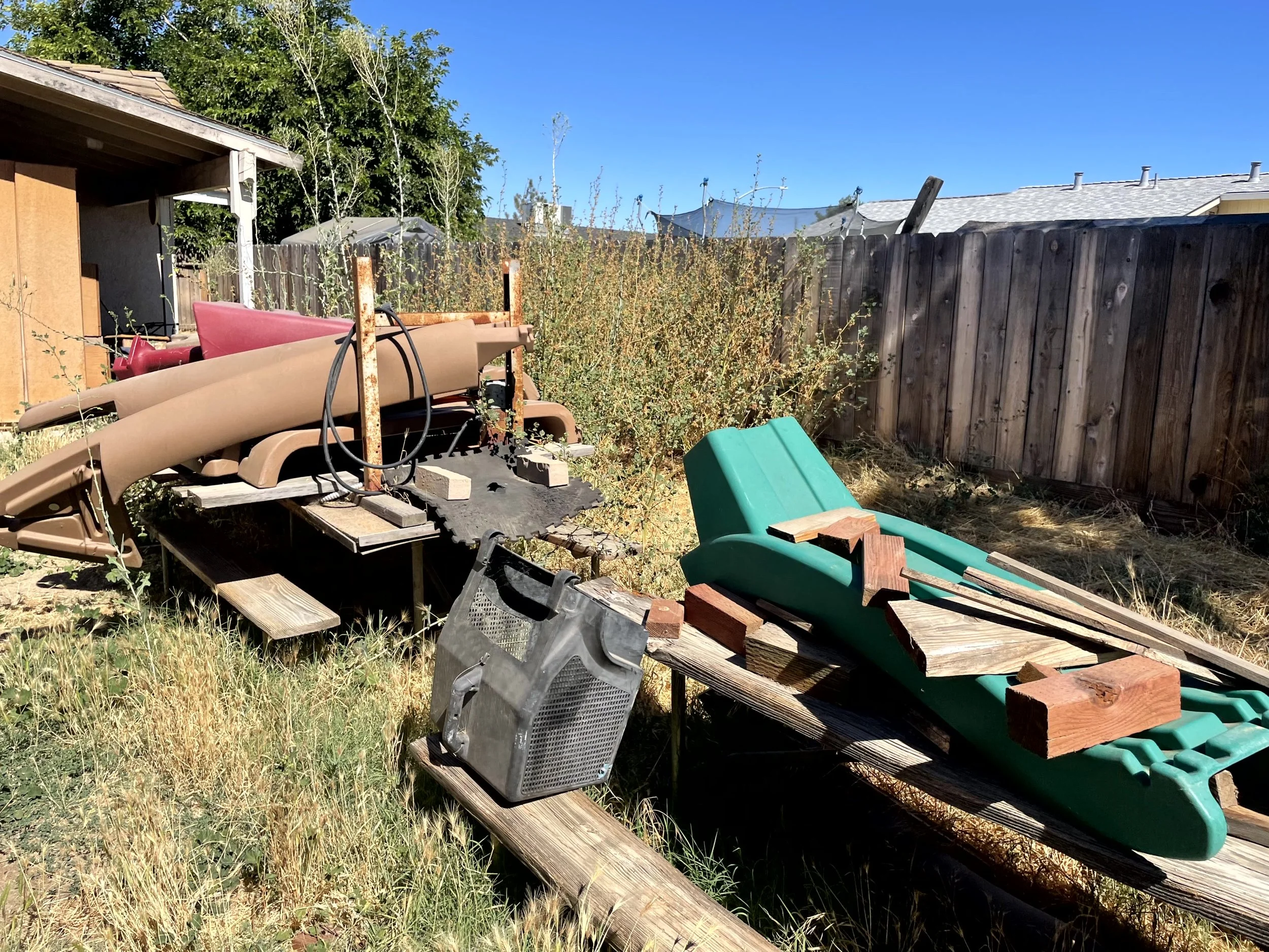 Backyard scene with abandoned lawn chairs, wooden planks, a plastic container, and overgrown weeds near a wooden fence on a sunny day.