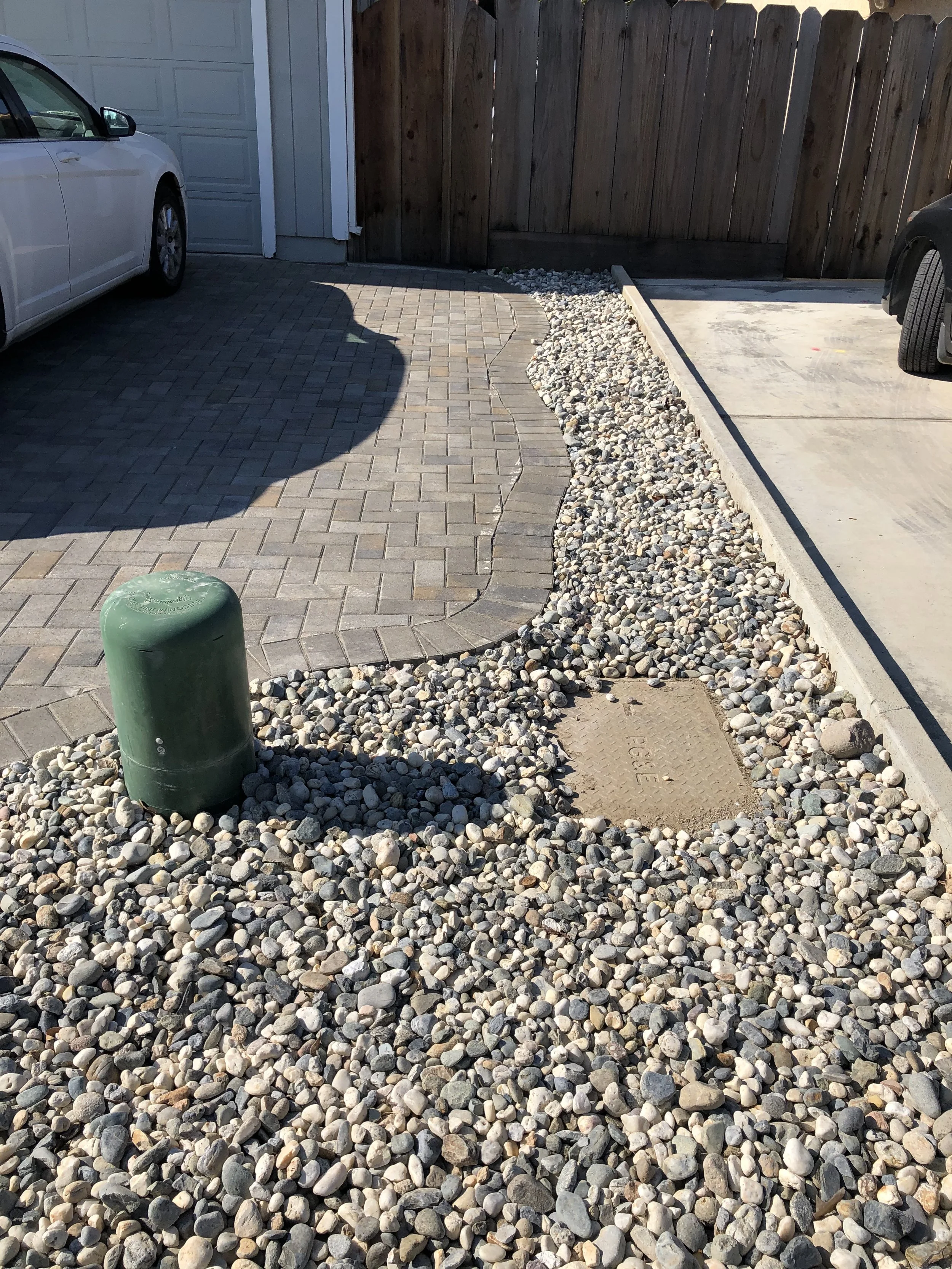 A driveway with a small paved section, surrounded by small rocks and a wooden fence. There are two parked cars, one on each side. A green bollard and a metal drain cover are also visible.