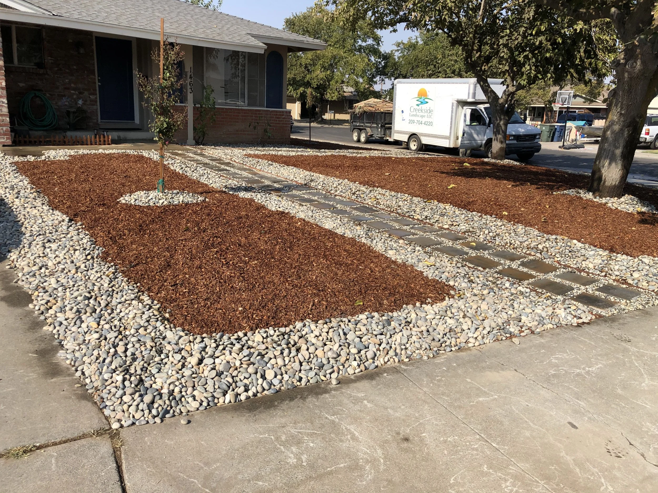 A landscaped front yard with two newly planted small trees, separated by a pathway with stepping stones. The yard is covered with reddish mulch and bordered with white decorative rocks, in front of a house with a porch.