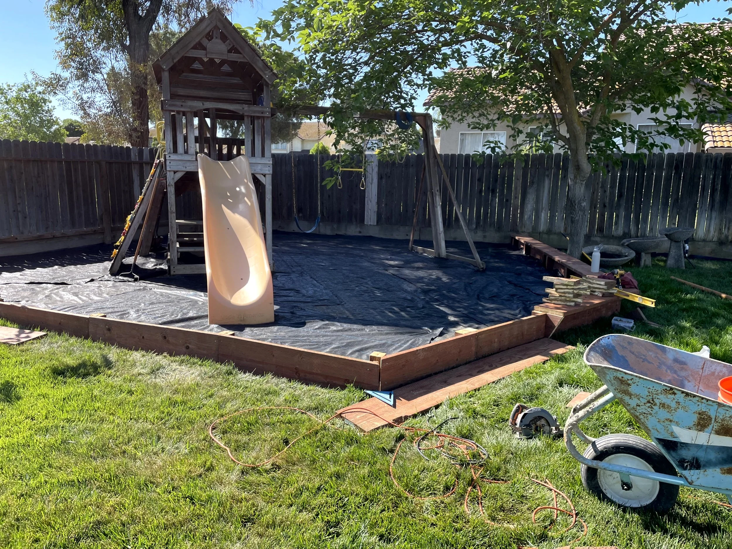 Backyard playground under construction with a wooden playset, slide, swings, and a sandbox on a black tarp, surrounded by green grass and a wooden fence.