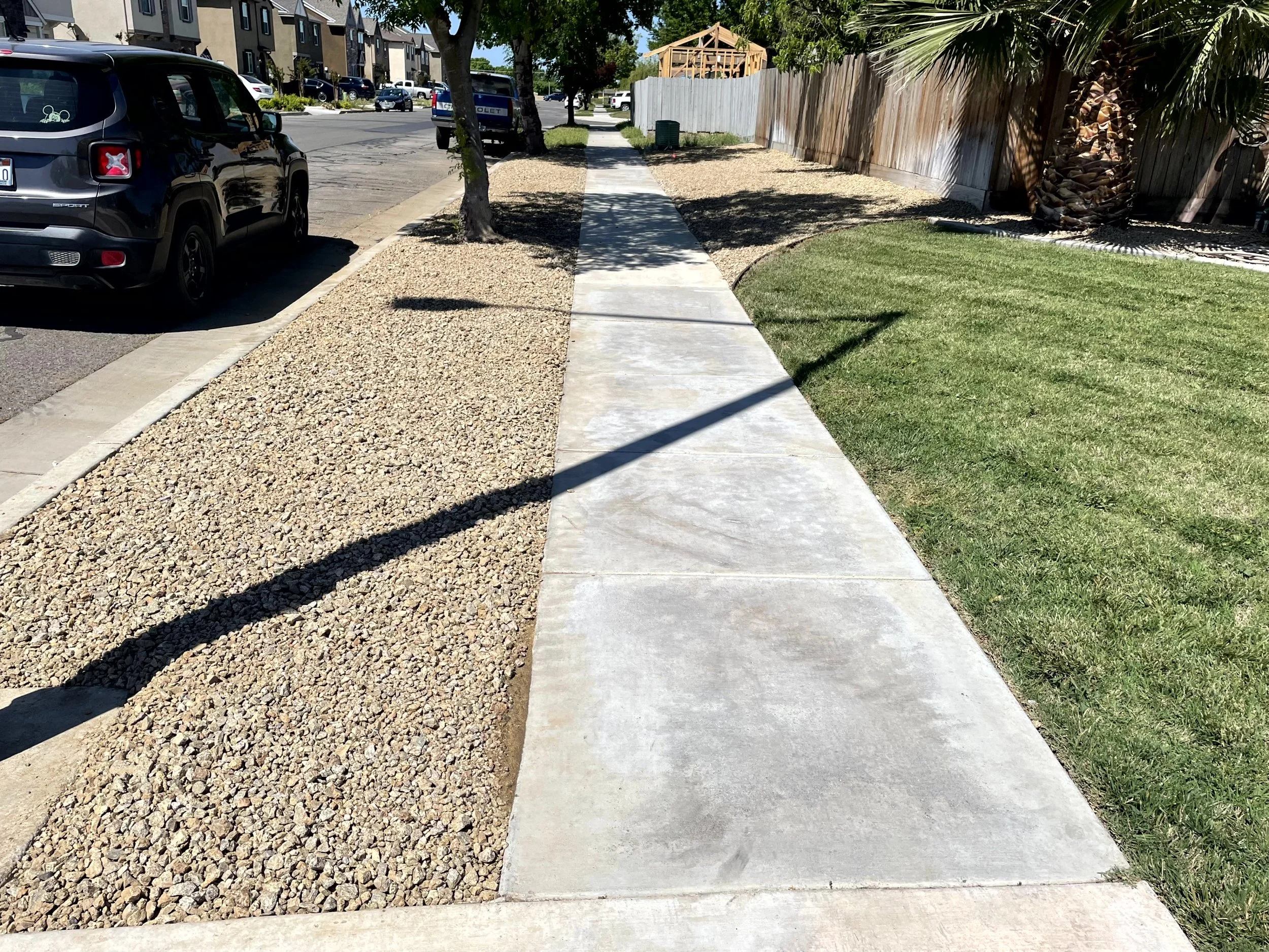 Sidewalk with concrete slabs, a grassy lawn, and a gravel area with a parked car and trees along a residential street.
