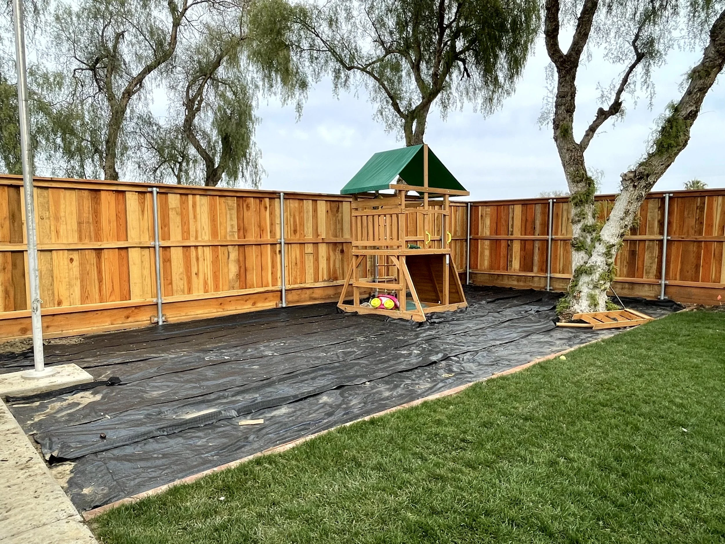 Backyard with a newly built wooden fence, a playhouse with a green roof, and a tree with a tire swing. The ground is covered with black landscaping fabric and some sports balls are under the playhouse.