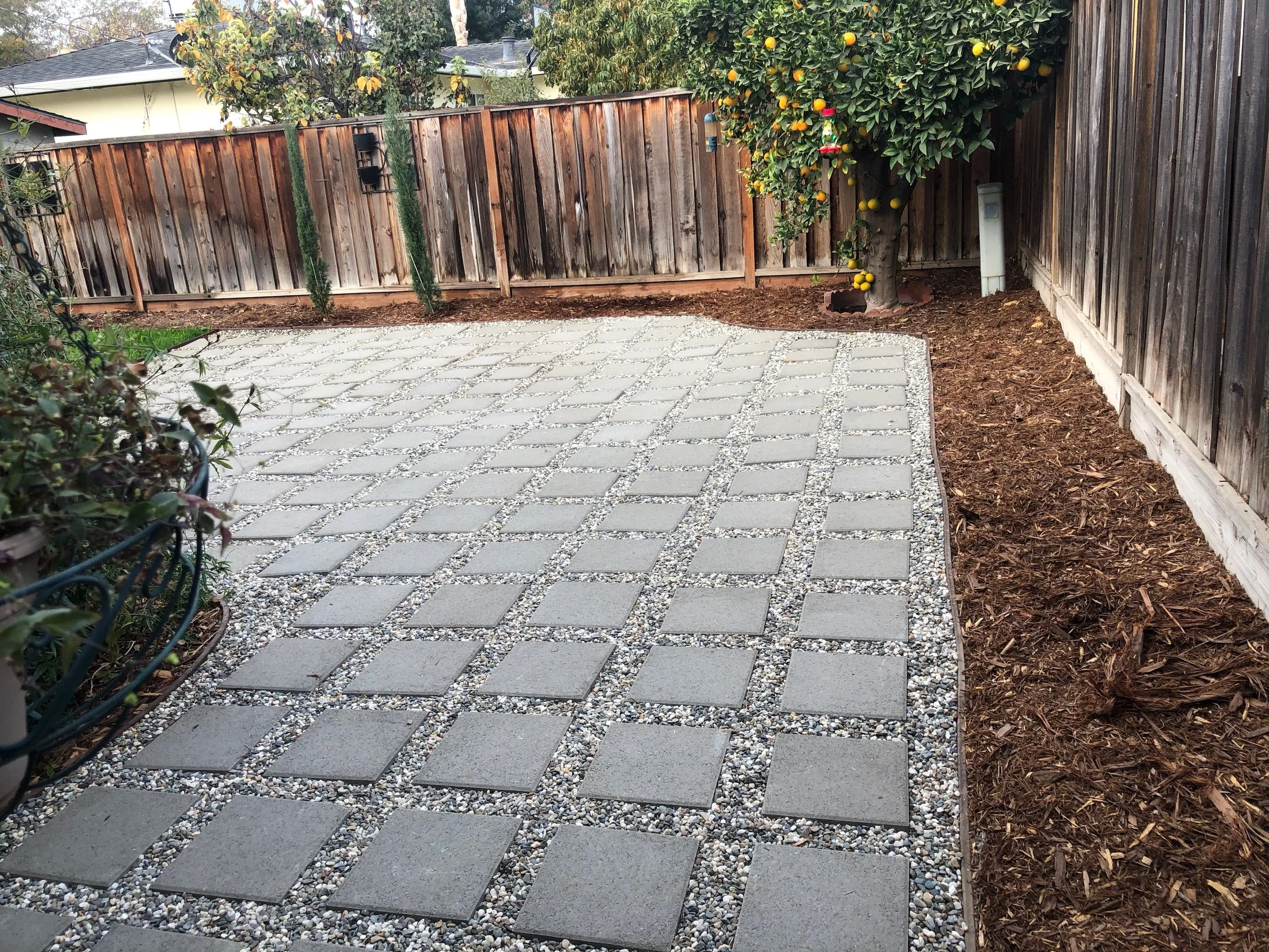A backyard patio with grey stone pavers arranged in a grid pattern, bordered by small white pebbles, with a wooden fence, a tree with yellow and red fruit, and some plants along the sides.