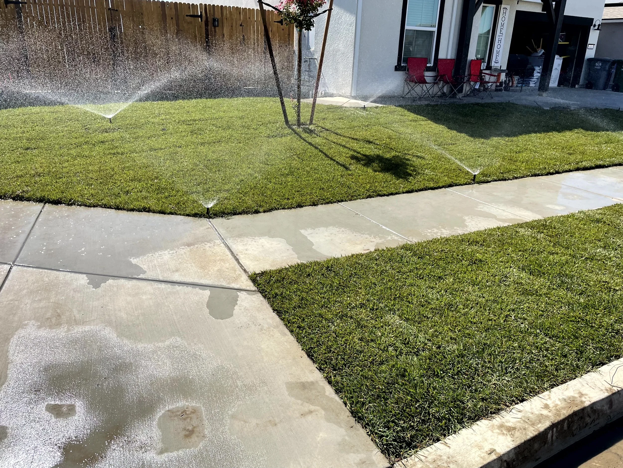 A residential lawn with newly patched grass, watering with sprinklers, and wet concrete sidewalk.