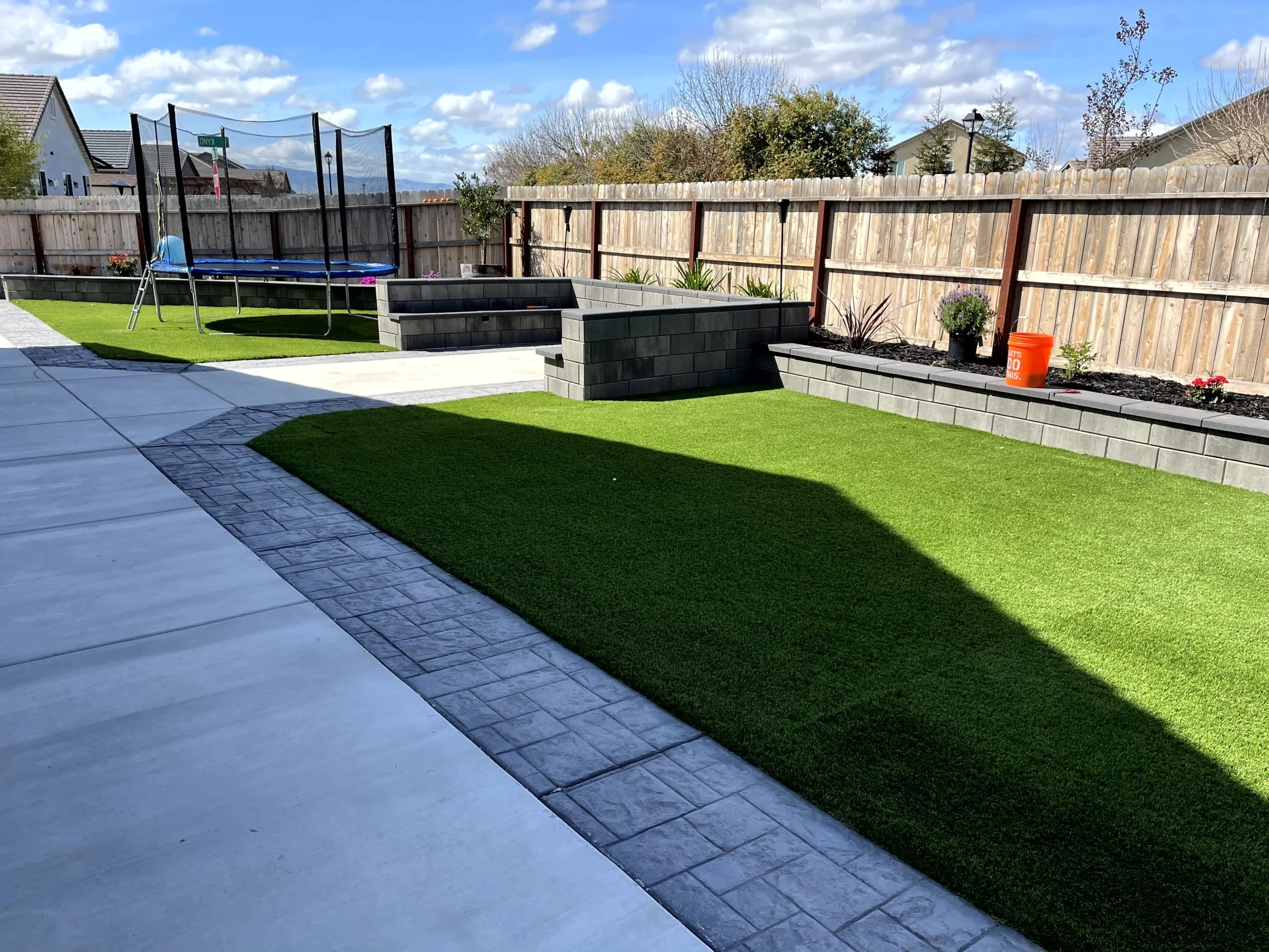Backyard with artificial grass, a trampoline, raised flower beds with plants, and a wooden privacy fence under a partly cloudy sky.