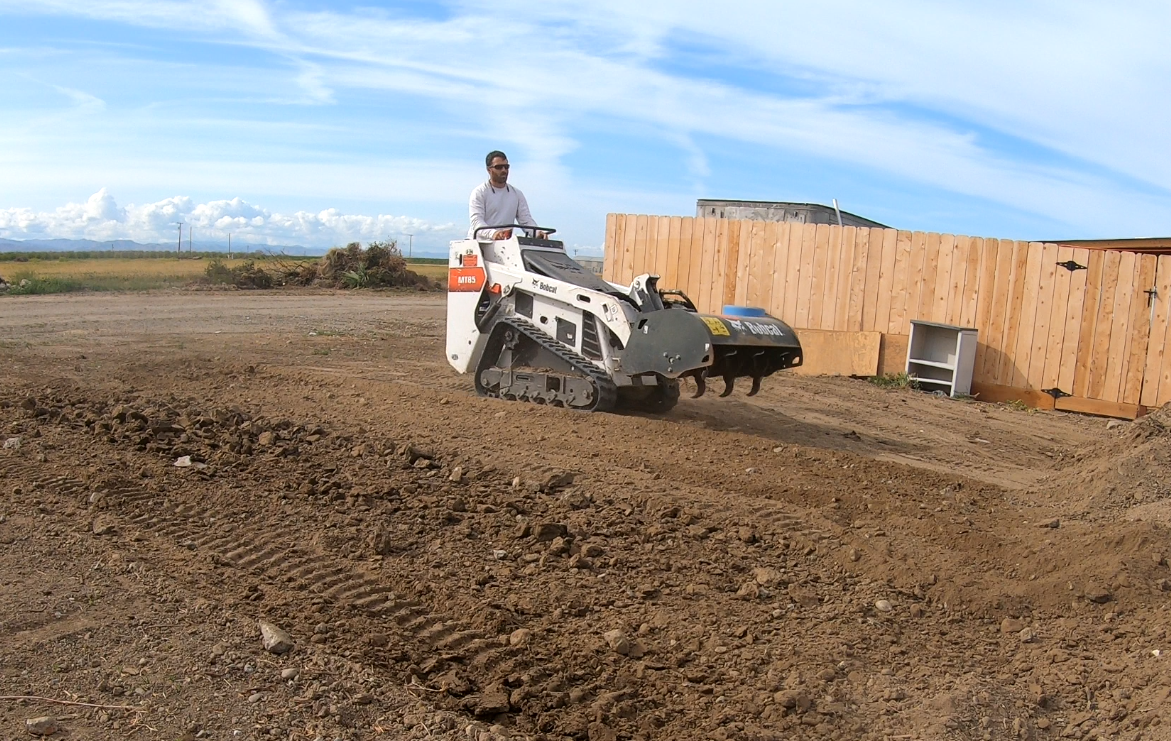A man operating a tracked mini bulldozer on a dirt lot with a wooden fence and open sky in the background.