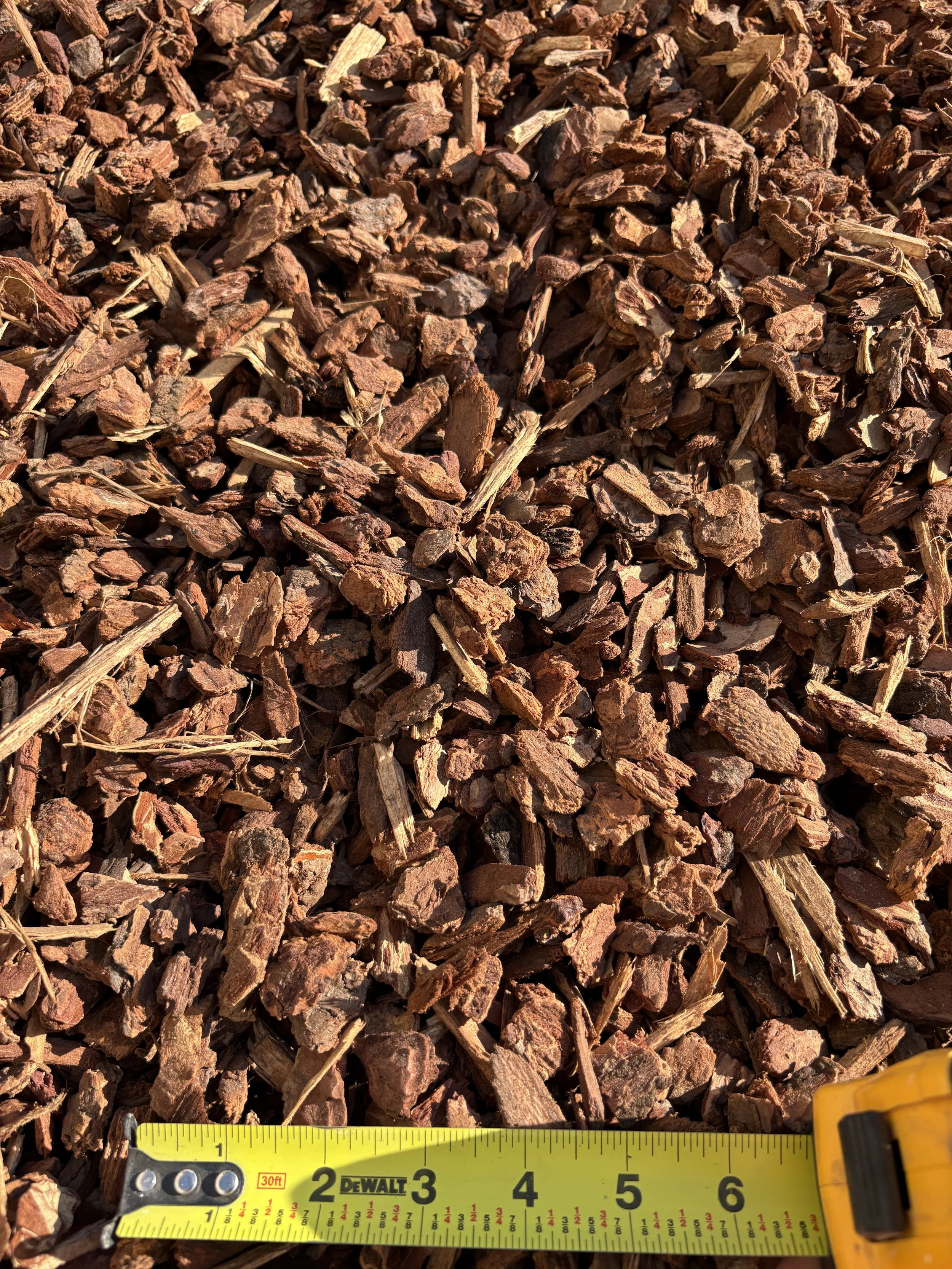 Close-up view of reddish-brown bark mulch with a yellow measuring tape showing around 6 inches in length at the bottom.