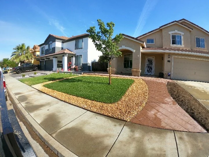 A suburban front yard with a young tree, green grass, and a gravel border. Two houses are visible, and the sky is clear with some wispy clouds.