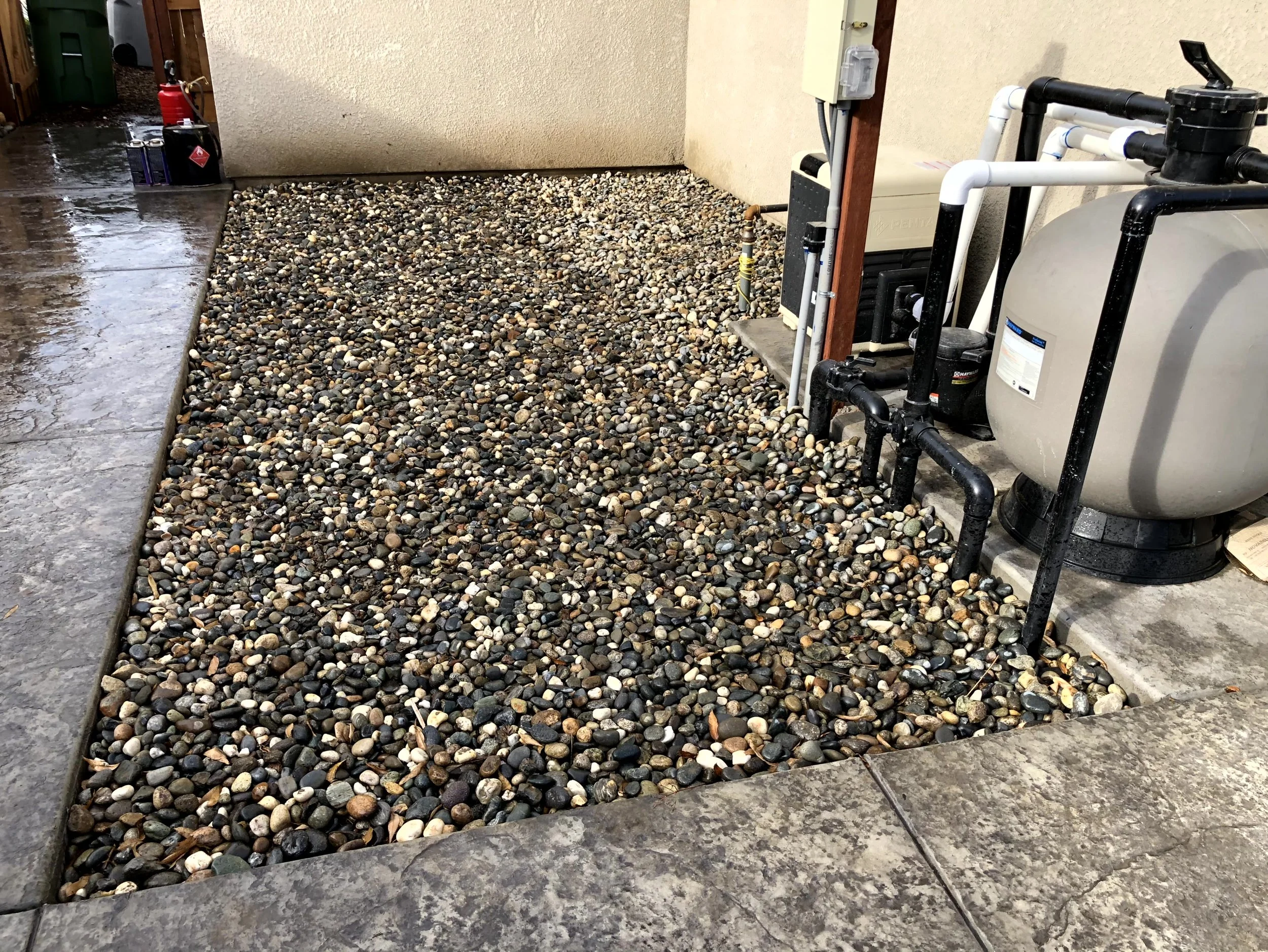 Section of outdoor yard with gravel and decorative stones next to a concrete patio, with some plumbing and equipment.