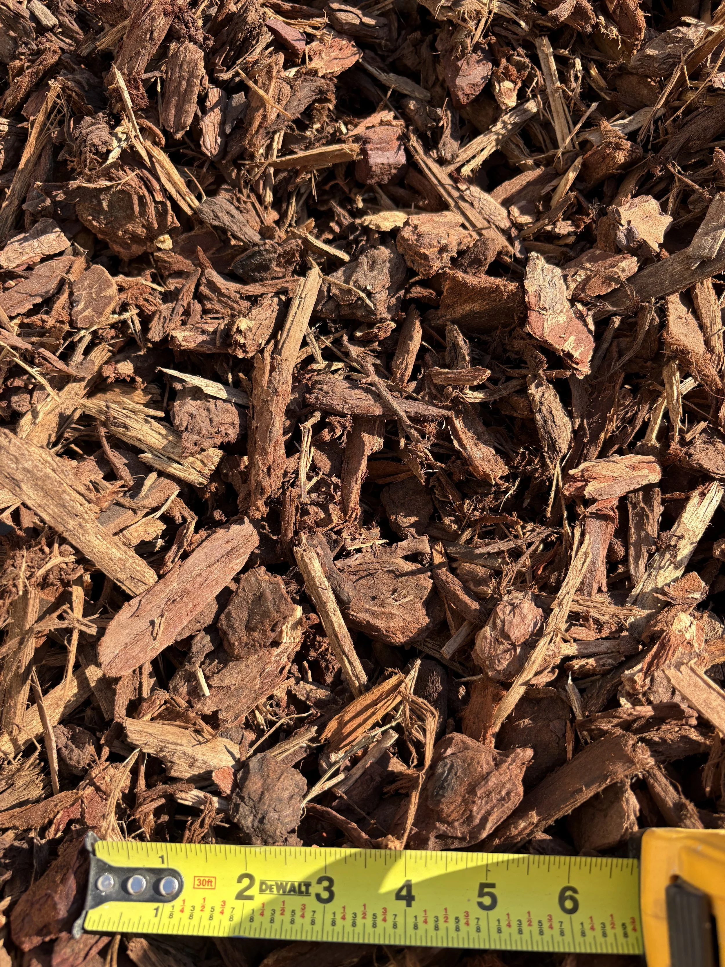Close-up of wood mulch with a yellow tape measure at the bottom showing measurements in inches.